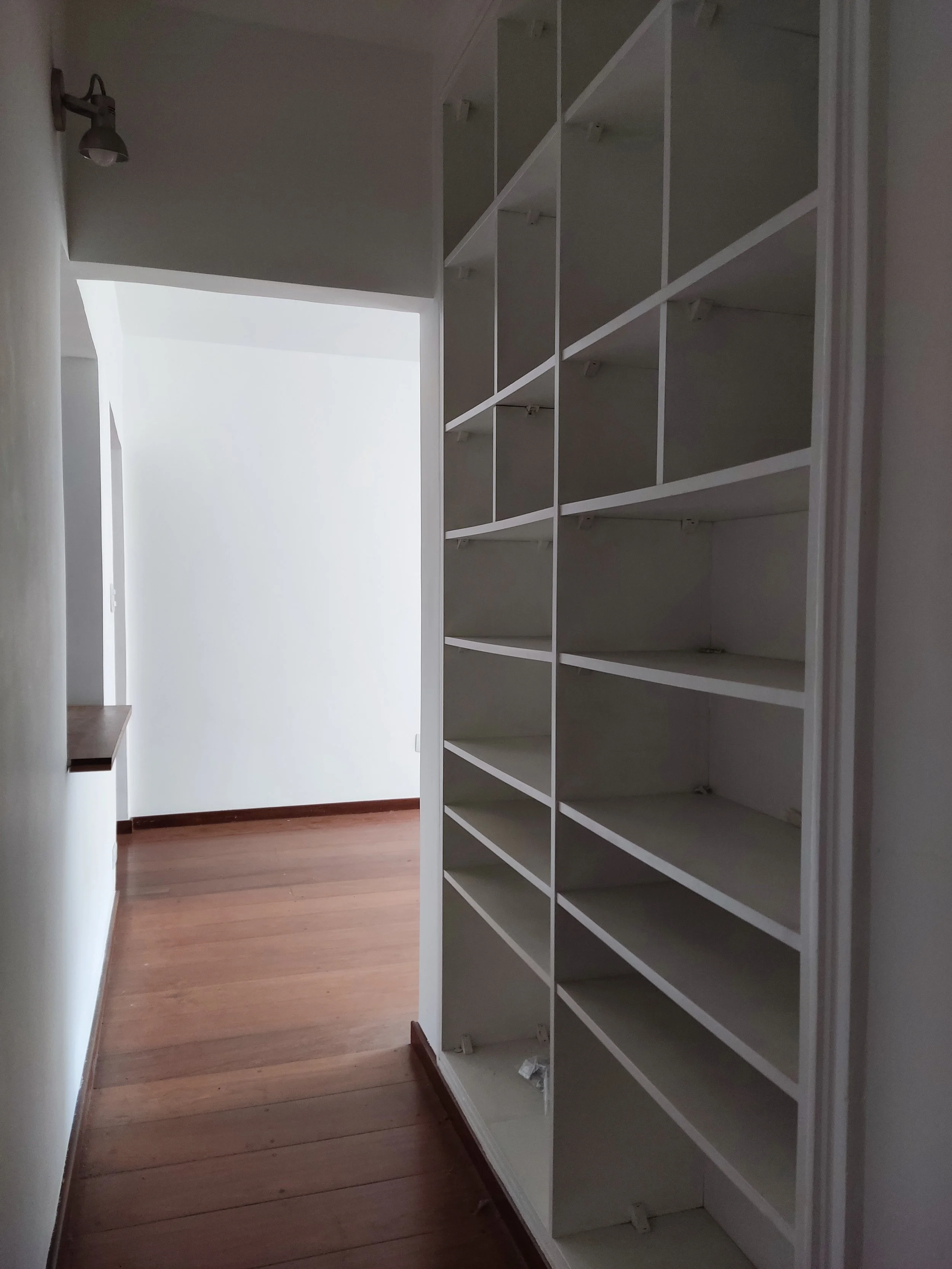 Corredor com prateleiras brancas na parede direita, piso de madeira, parede branca ao fundo e iluminação de spot no teto esquerdo.