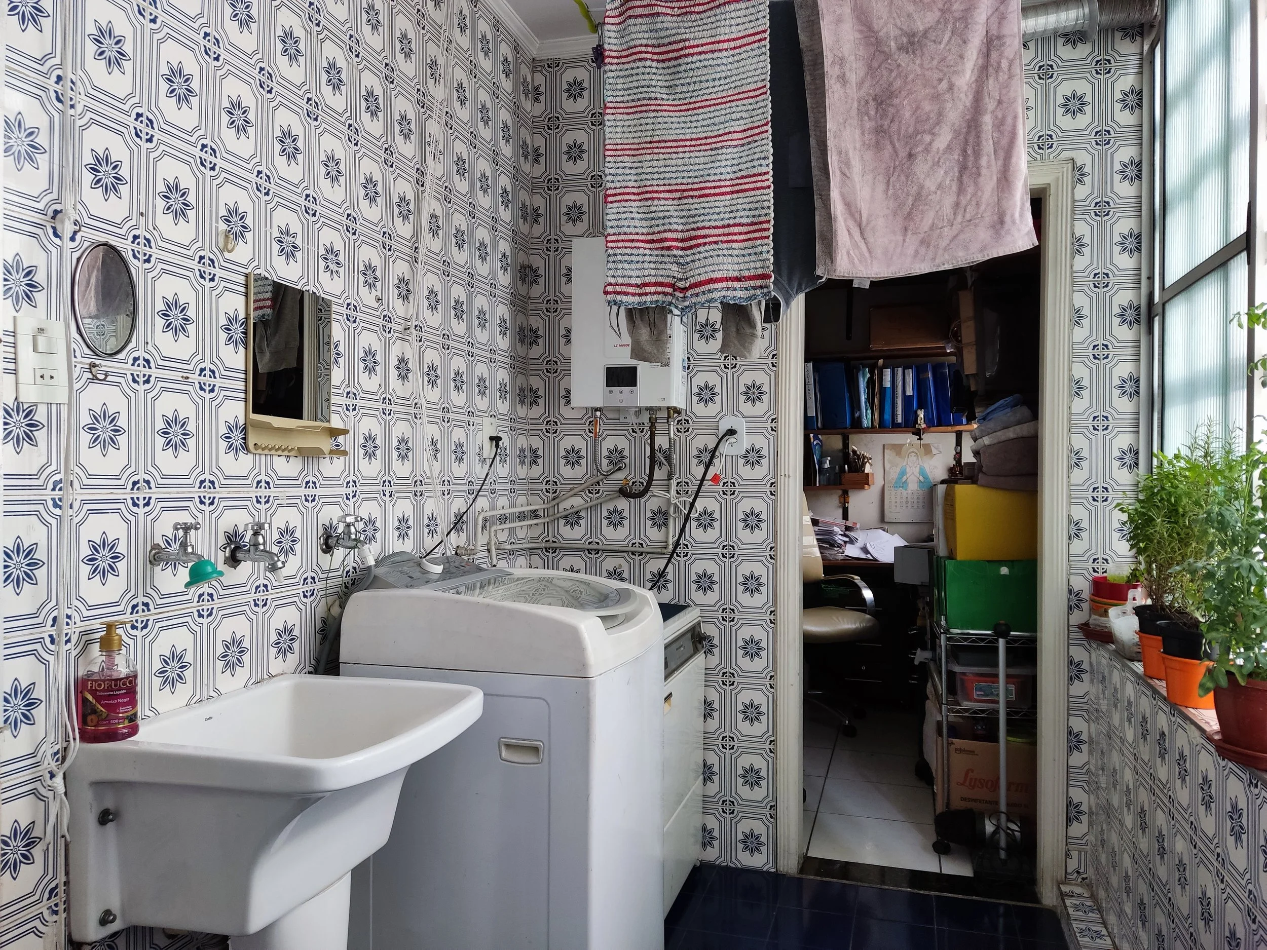 Lavanderia com parede com azulejos decorativos azuis e brancos, pia branca, lavabo, máquina de lavar roupas e uma área de varal com roupas penduradas. Ao fundo, uma porta leva a um escritório pequeno com várias prateleiras, papéis, livros, janelas de