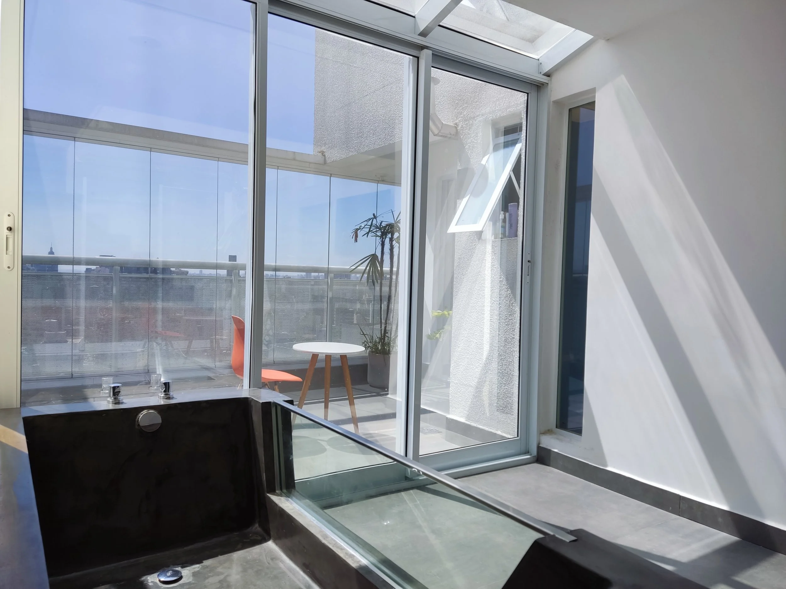 Sala de banho com banheira preta e vidro do lado, área externa com mesa branca, cadeira laranja e plantas, vista do céu limpo.