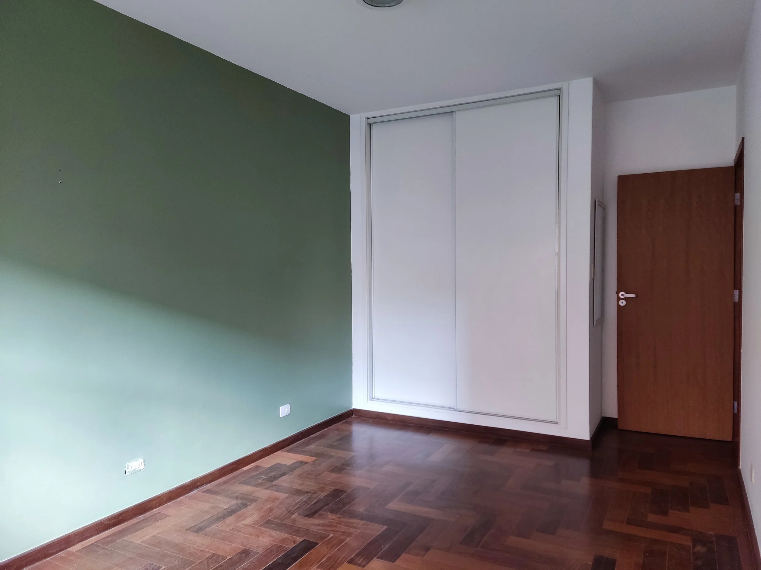 Quarto sem móveis com parede verde na esquerda, parede branca na direita e um armário embutido branco com portas de correr, além de uma porta de madeira opaca ao lado do armário.