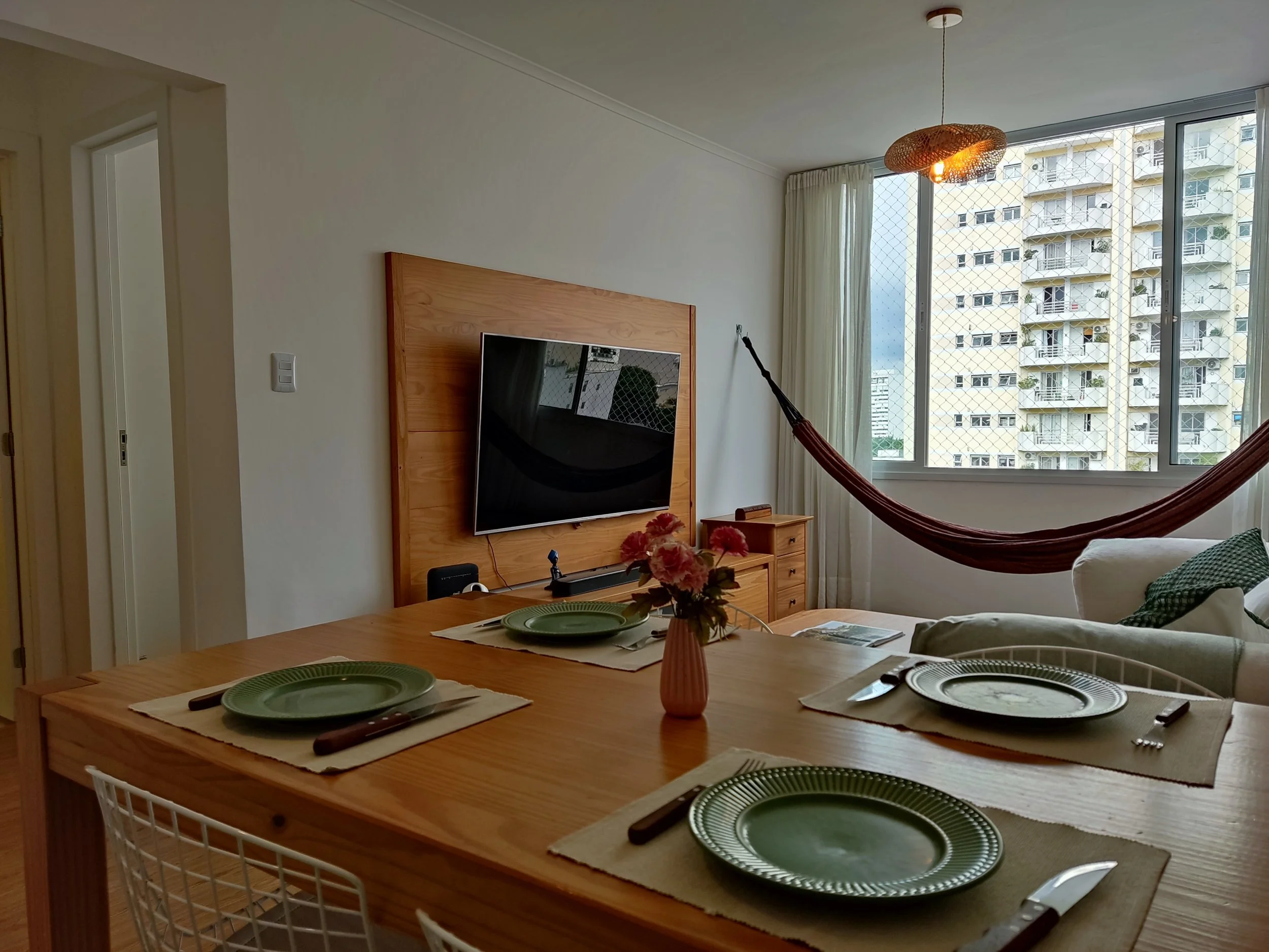 Sala de estar com mesa de jantar de madeira, quatro pratos verdes, talheres, vaso com flores, sofá branco com almofada verde, rede de descanso perto da janela grande com vista para edifícios altos, cortinas brancas, luminária de teto de fibra.