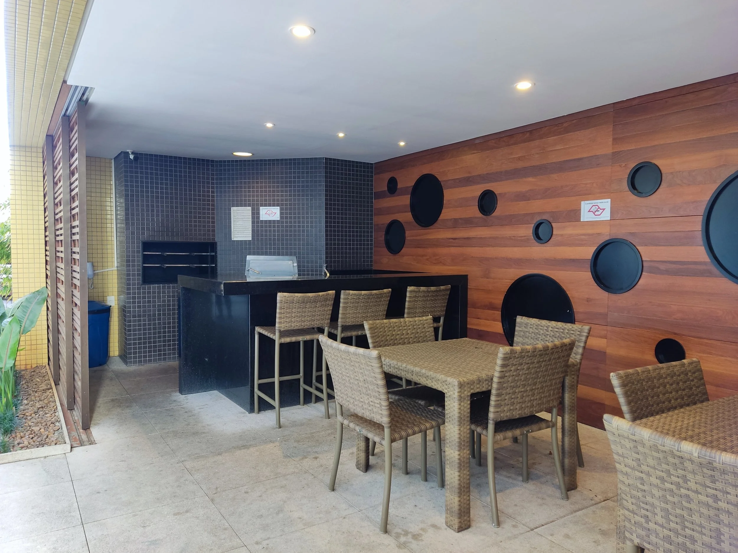 Espaço de convivência com mesa de madeira, cadeiras de fibras, parede de madeira com círculos pretos decorativos, balcão de atendimento, parede com azulejos pretos e área externa com plantas.