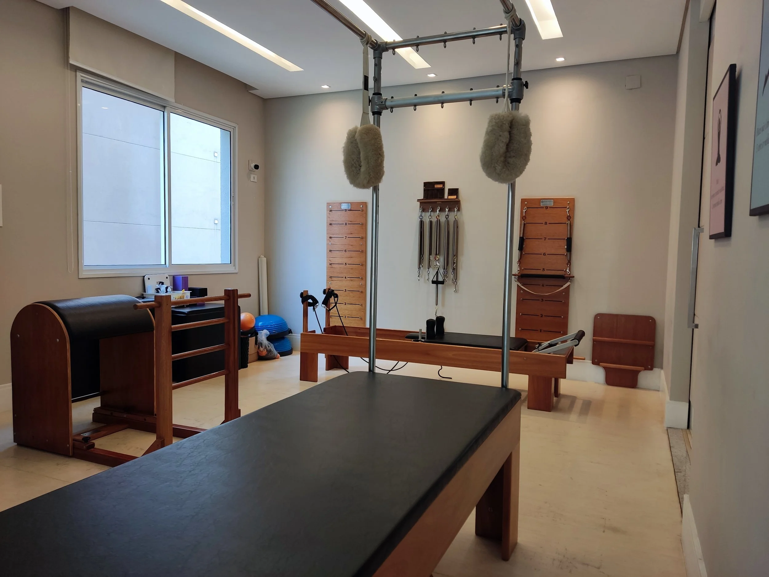 Sala de fisioterapia com equipamentos como estiramento, bolas, lycra, travessas e aparelho de pilates.