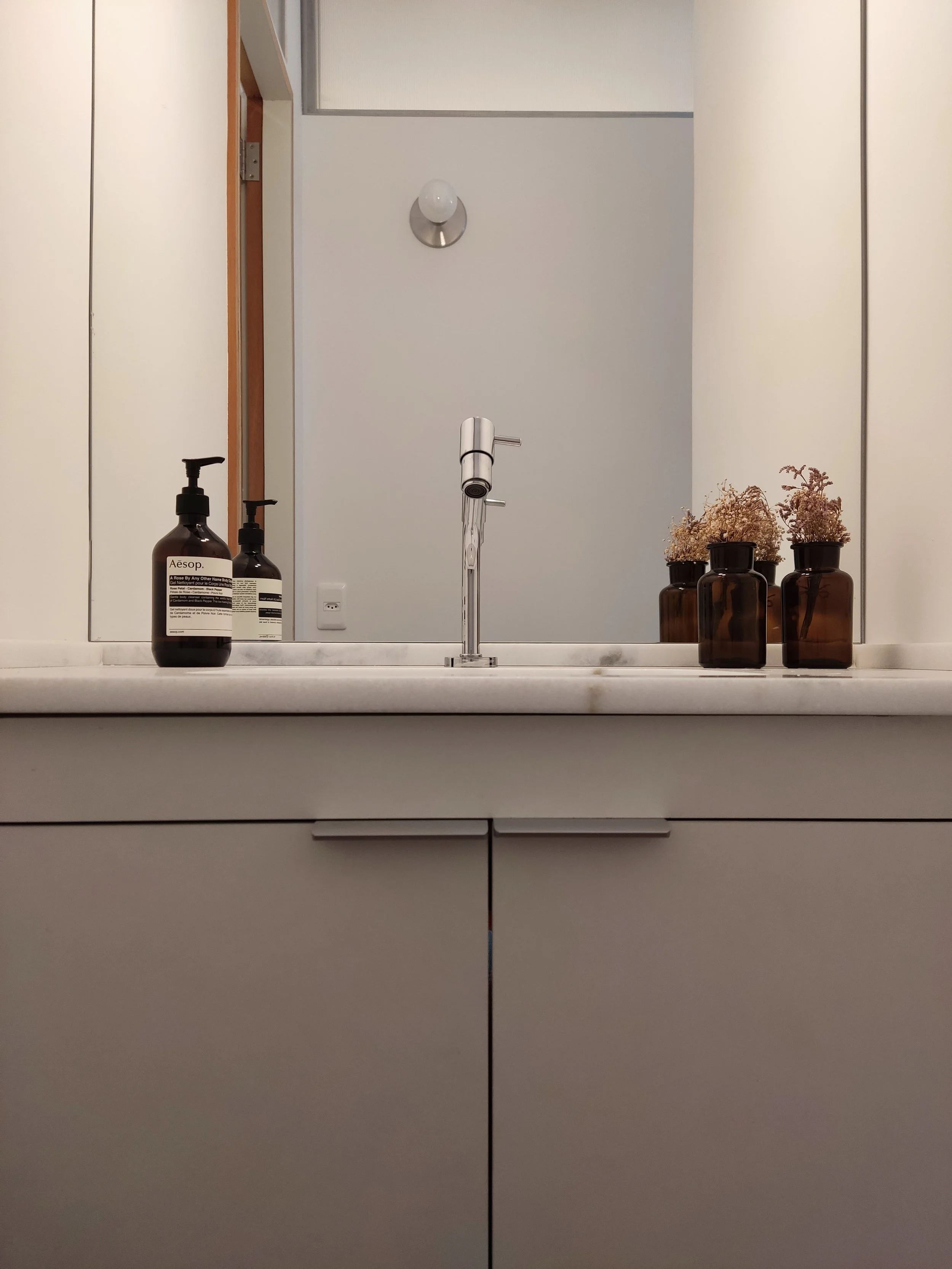 Espelho acima de uma pia com sabonete líquido e três frascos decorativos na bancada.