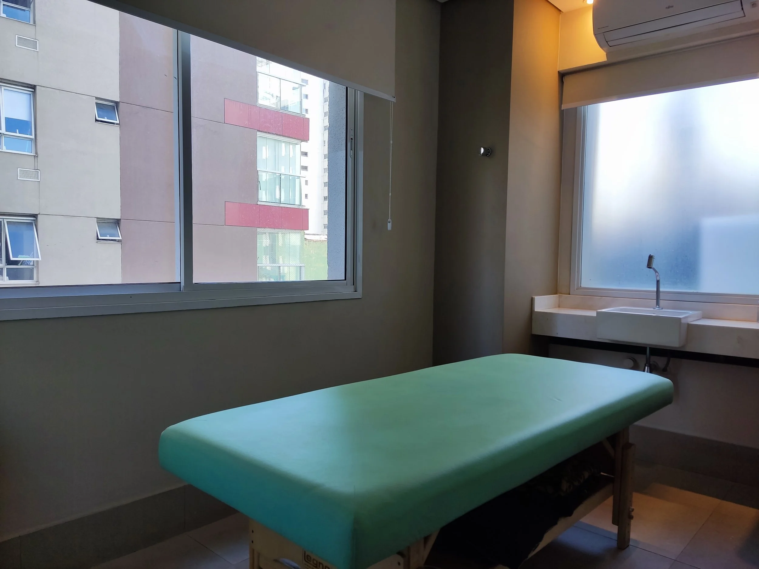 Sala de exame médica com mesa de exame verde, janelas grandes com vista para prédios, pia junto à janela ao fundo, no interior de consultório.