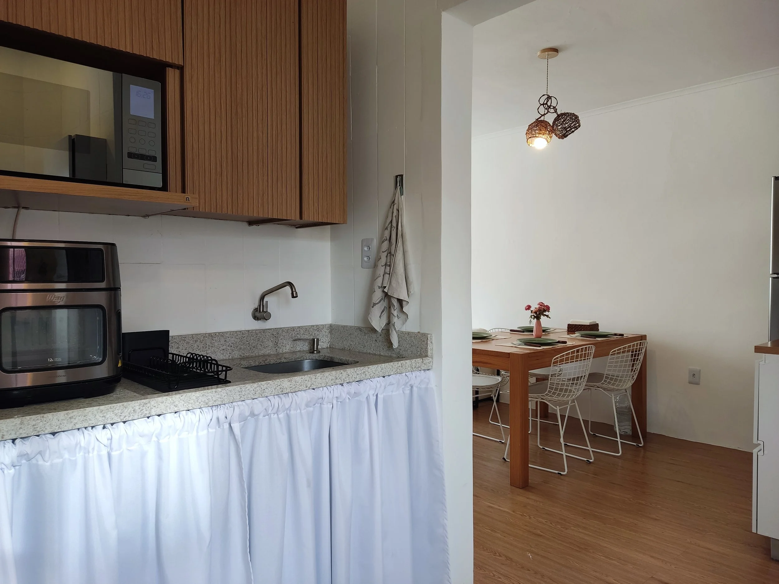 Cozinha com armários de madeira, micro-ondas embutido, forno elétrico, pia com torneira, pia de inox, utensílios de cozinha, cortina branca na base do balcão, e uma sala de jantar ao fundo com mesa de madeira, cinco cadeiras brancas, uma floricultura