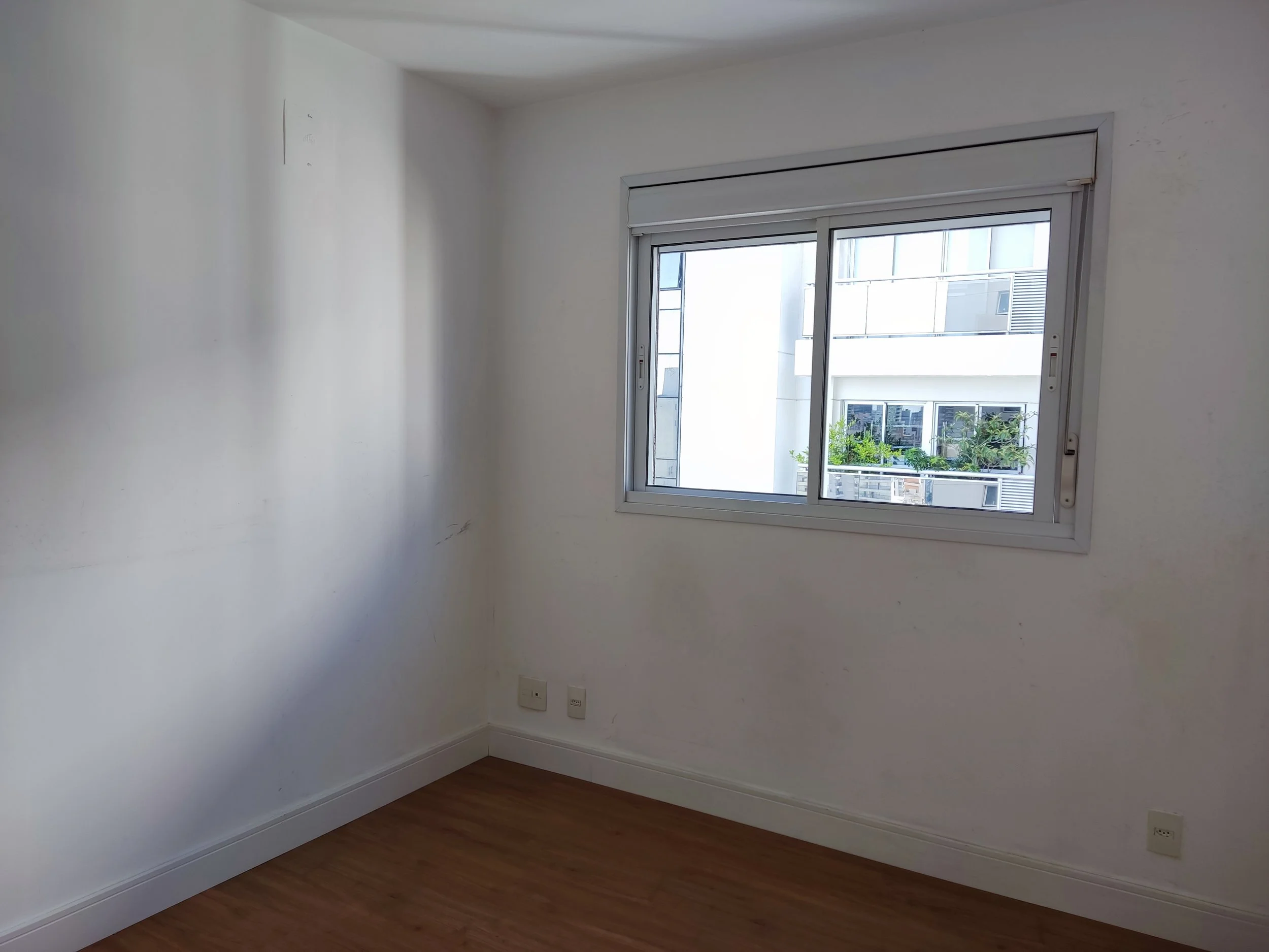 Quarto vazio com piso de madeira, parede branca, janela de correr com vista para prédios e plantas, fiação e tomadas na parede.