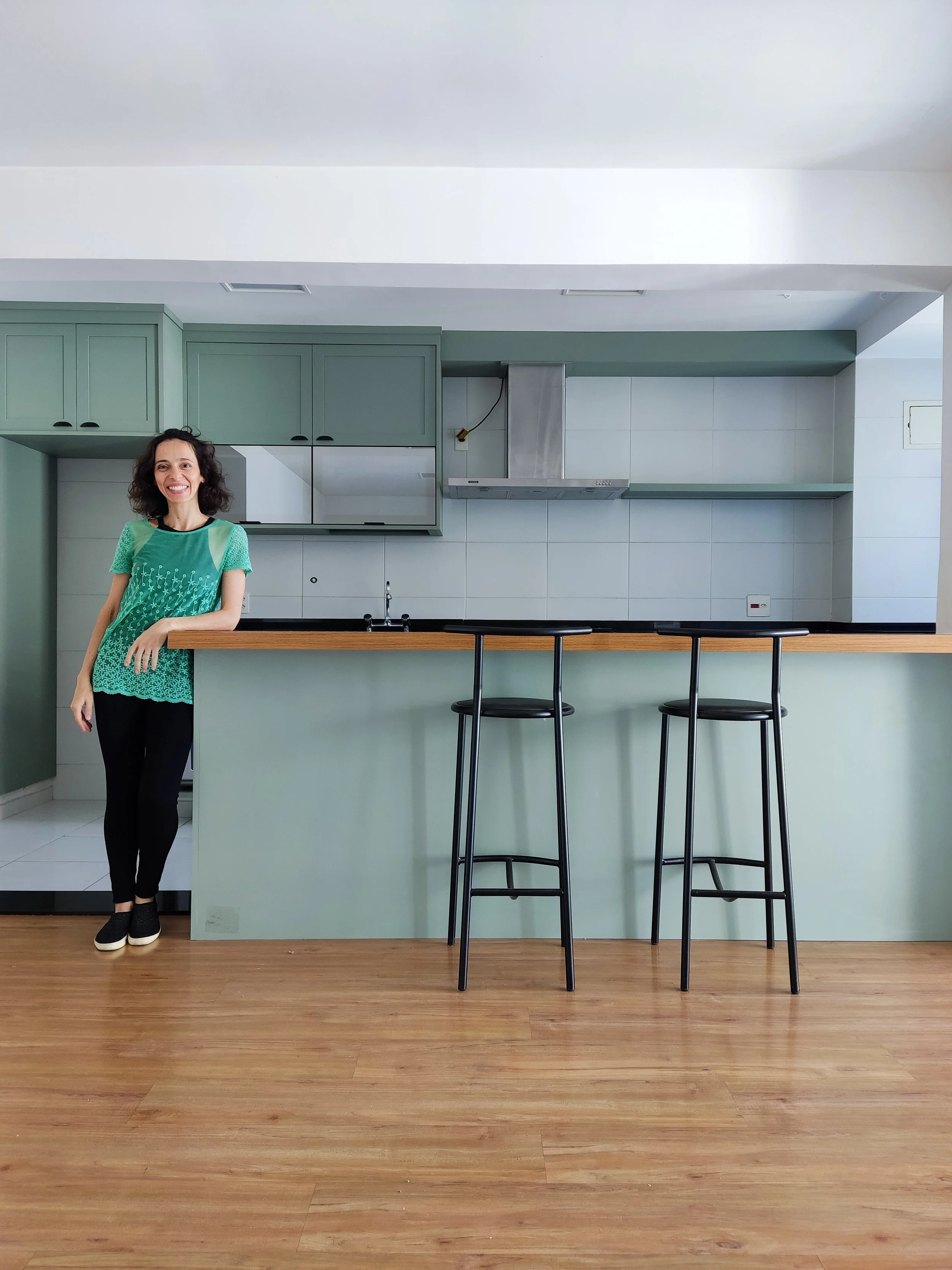 Mulher sorridente de cabelo cacheado, usando blusa verde, apoiada em balcão de cozinha moderna com cadeiras altos ao frente. Cozinha com armários verdes claros e eletrodomésticos de aço inox.