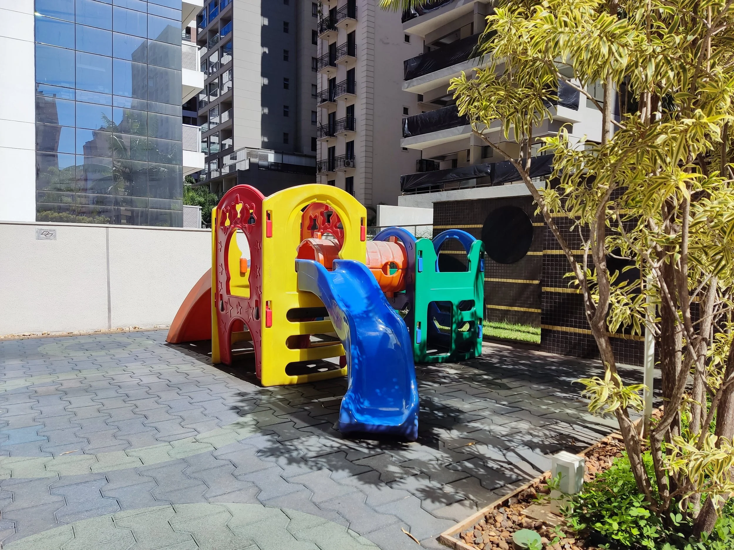Playground infantil com escorregador colorido em uma área ao ar livre, cercada por árvores e edifícios modernos ao fundo.