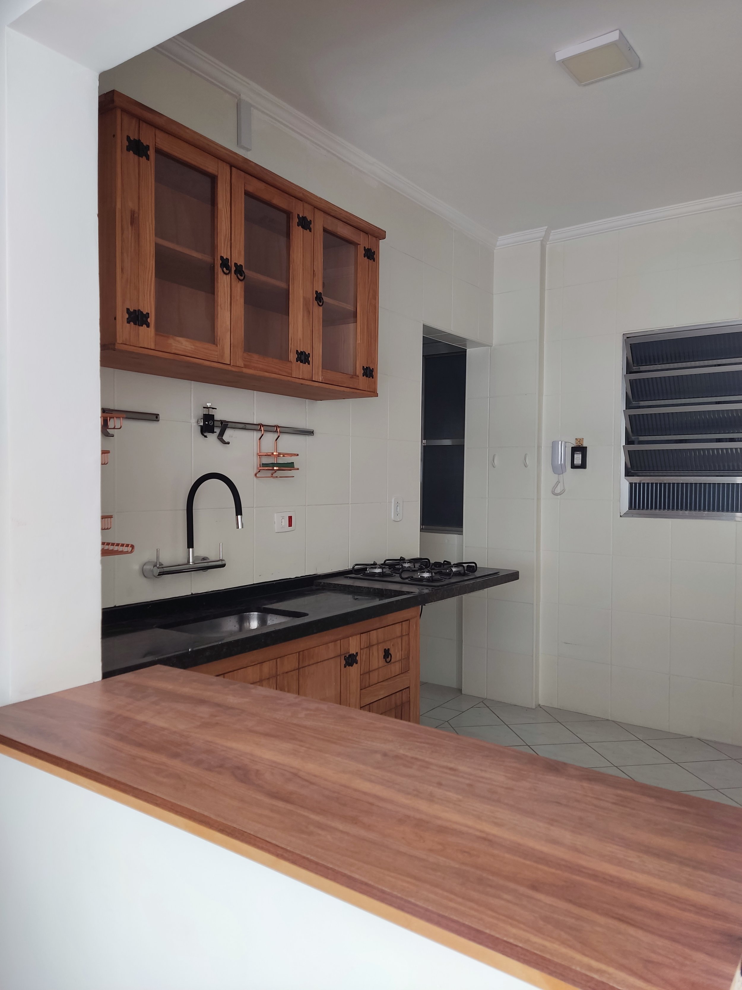 Cozinha moderna com bancada de madeira, armários de madeira, pia preta, fogão à gás, janela com persianas, interfone na parede e utensílios de cozinha prendidos em uma barra.