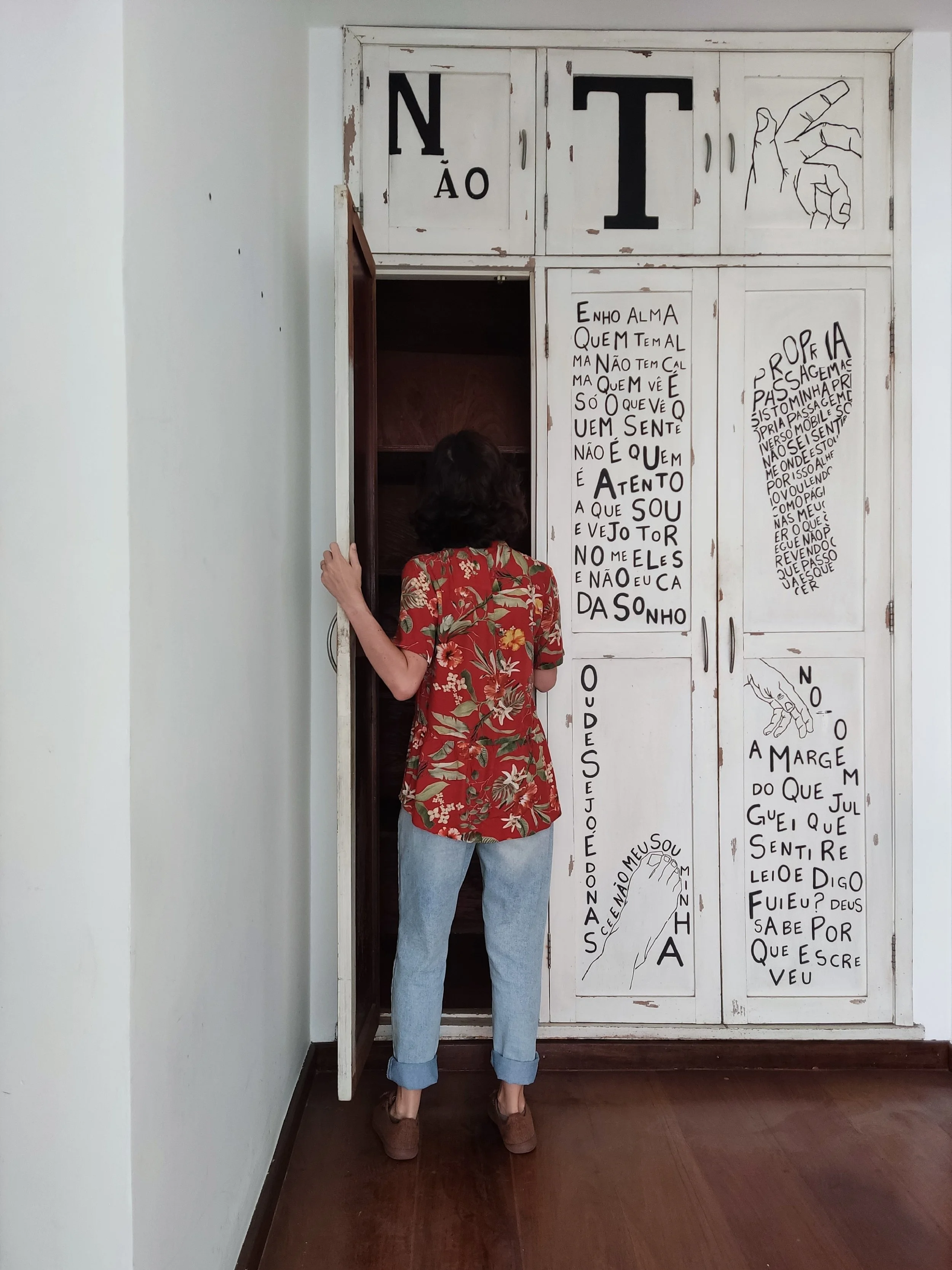 Pessoa de costas, usando uma blusa floral vermelha com costas nuas e calças jeans, entra em um armário antigo de portas branqueadas com textos escritos à mão nas portas.