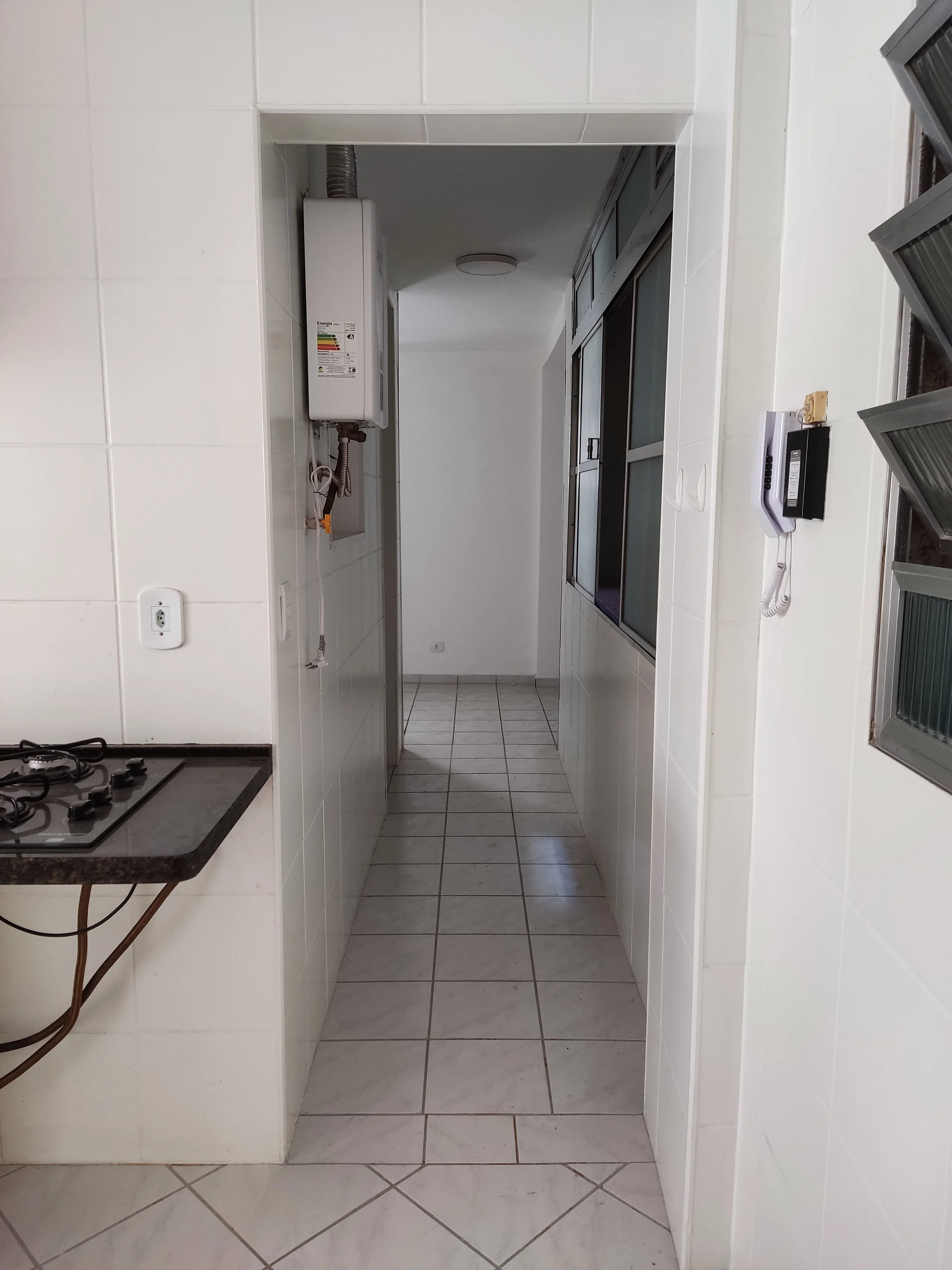 Corredor de uma cozinha com piso de azulejo branco, paredes azulejadas brancas, com janela de vidro e aparelho de telefone na parede à direita. Fila de armários ao lado direito e uma porta ao fundo.