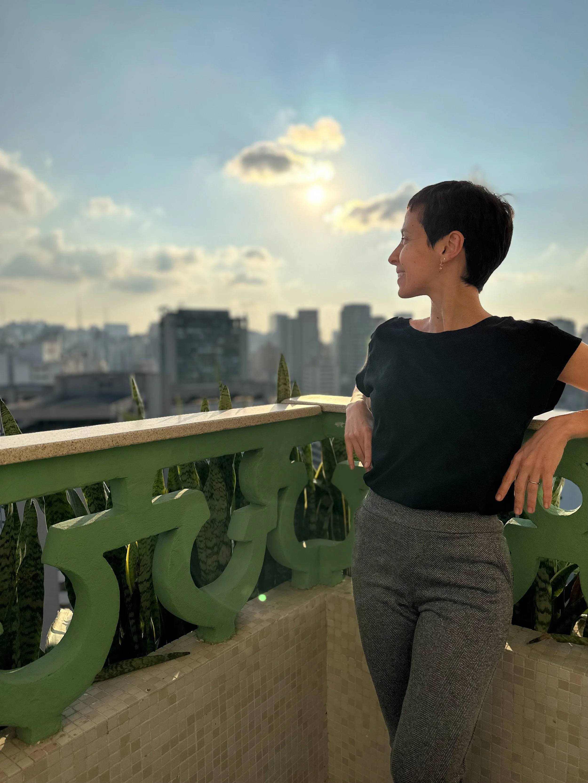Mulher com cabelo curto, vestindo blusa preta e calça de pantalona, se apoia na borda de um terraço com paisagem urbana ao fundo, durante o pôr do sol.