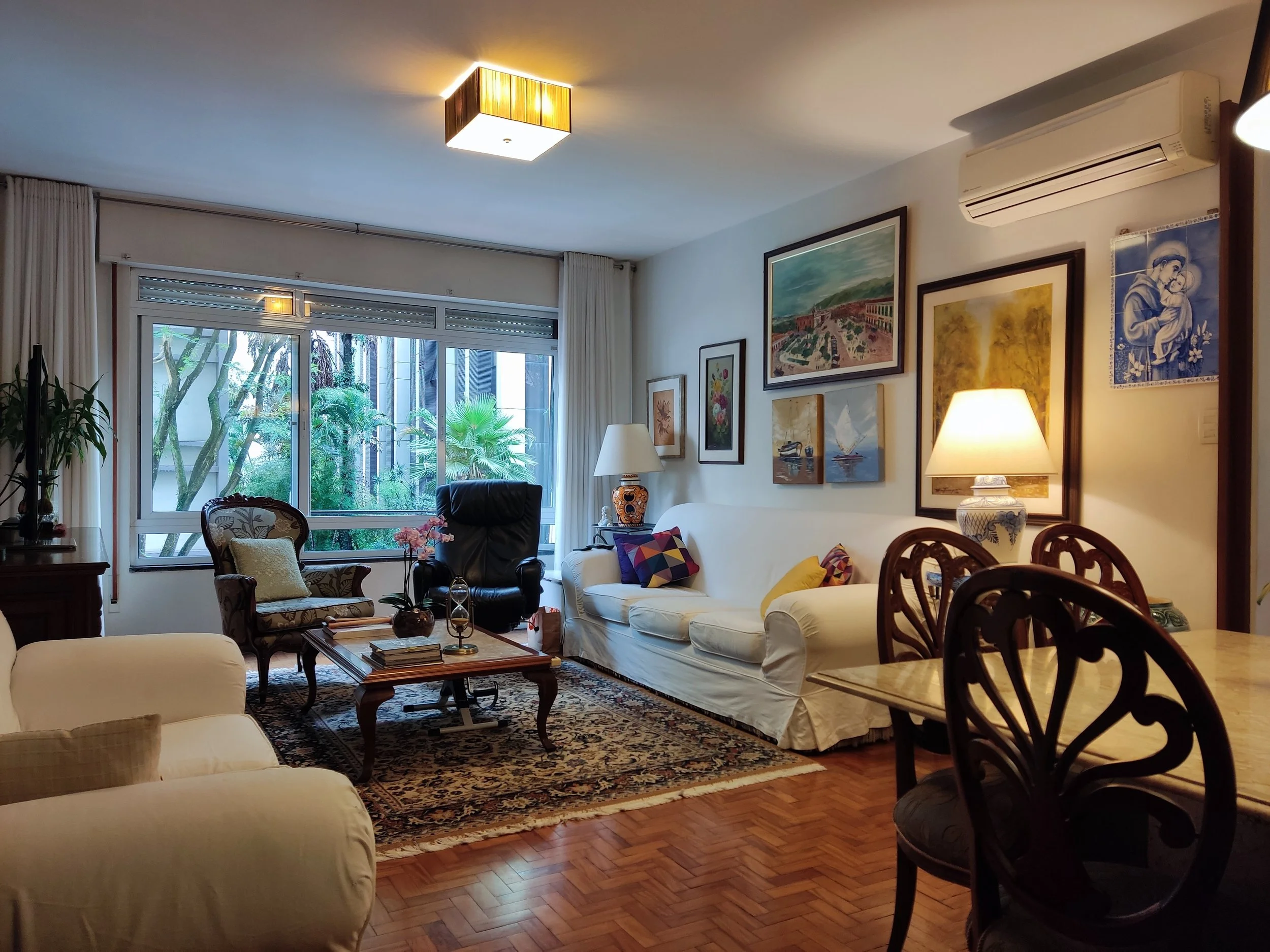 Sala de estar decorada com quadros nas paredes, sofá branco, poltrona, mesa de centro, luminárias, cortinas e janelas com vista para um jardim