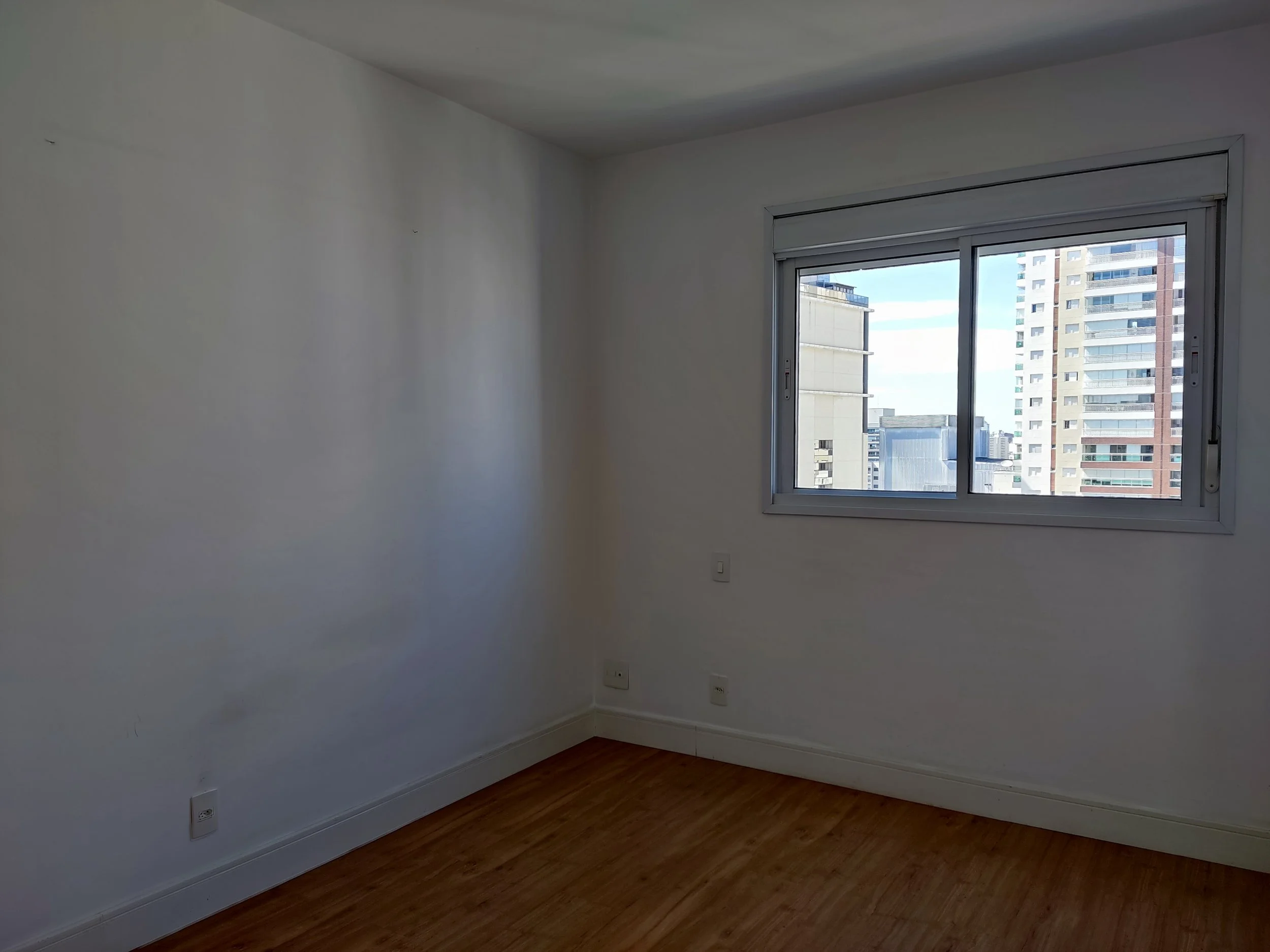 Quarto vazio com parede branca, janela grande com vista de prédios altos, piso de madeira clara