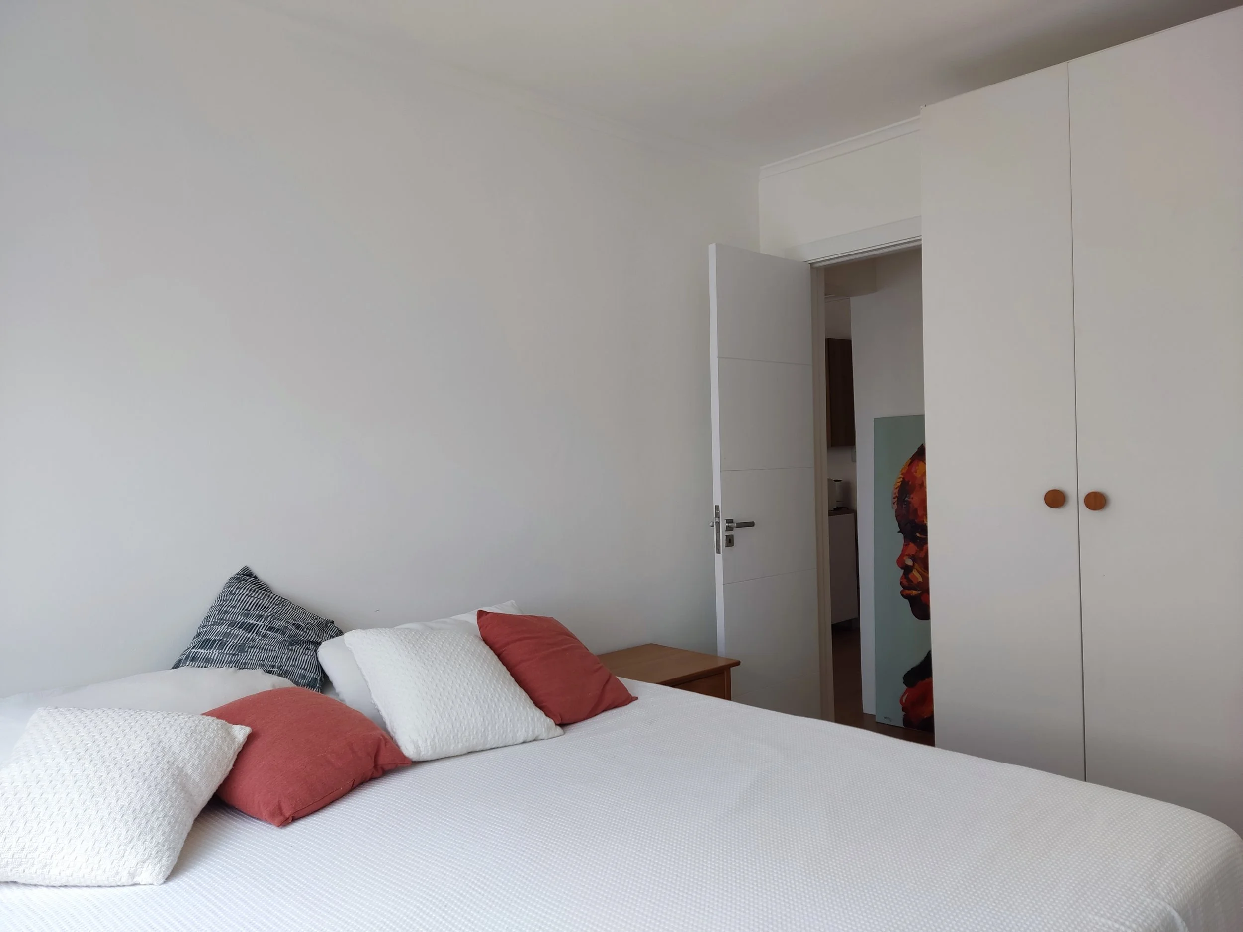 Quarto com cama de colchão branco, várias almofadas em diferentes cores e texturas ao lado, armário branco com portas de madeira ao lado, e porta aberta mostrando um corredor com uma obra de arte de rosto humano na parede.
