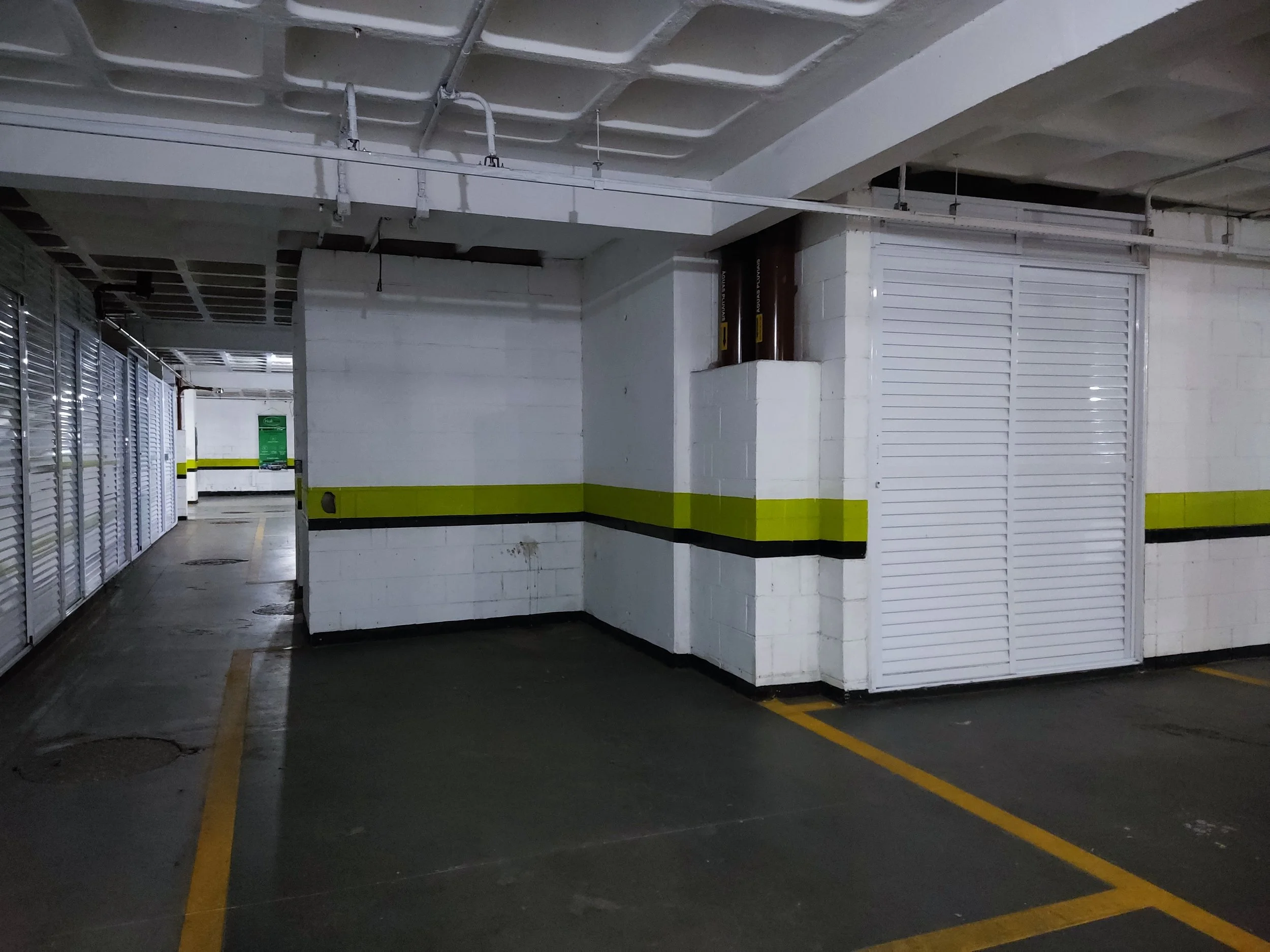 Estacionamento subterrâneo vazio com paredes brancas, faixa amarela e preta, portões de metal brancos e marcações de vagas no chão.