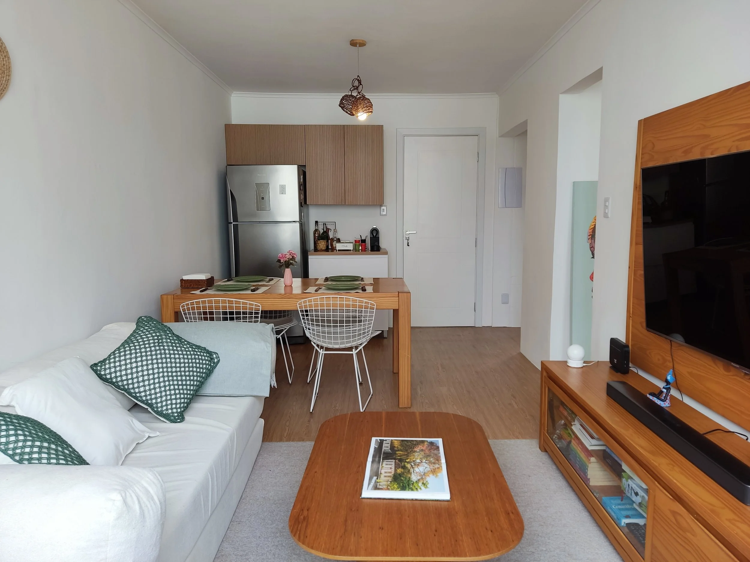 Sala de estar com sofá branco, almofadas verdes, mesa de centro de madeira, televisão em suporte de madeira, mesa de jantar de madeira com quatro cadeiras, cozinha com geladeira de inox e armários de madeira, decoração com vaso de flores e objetos di