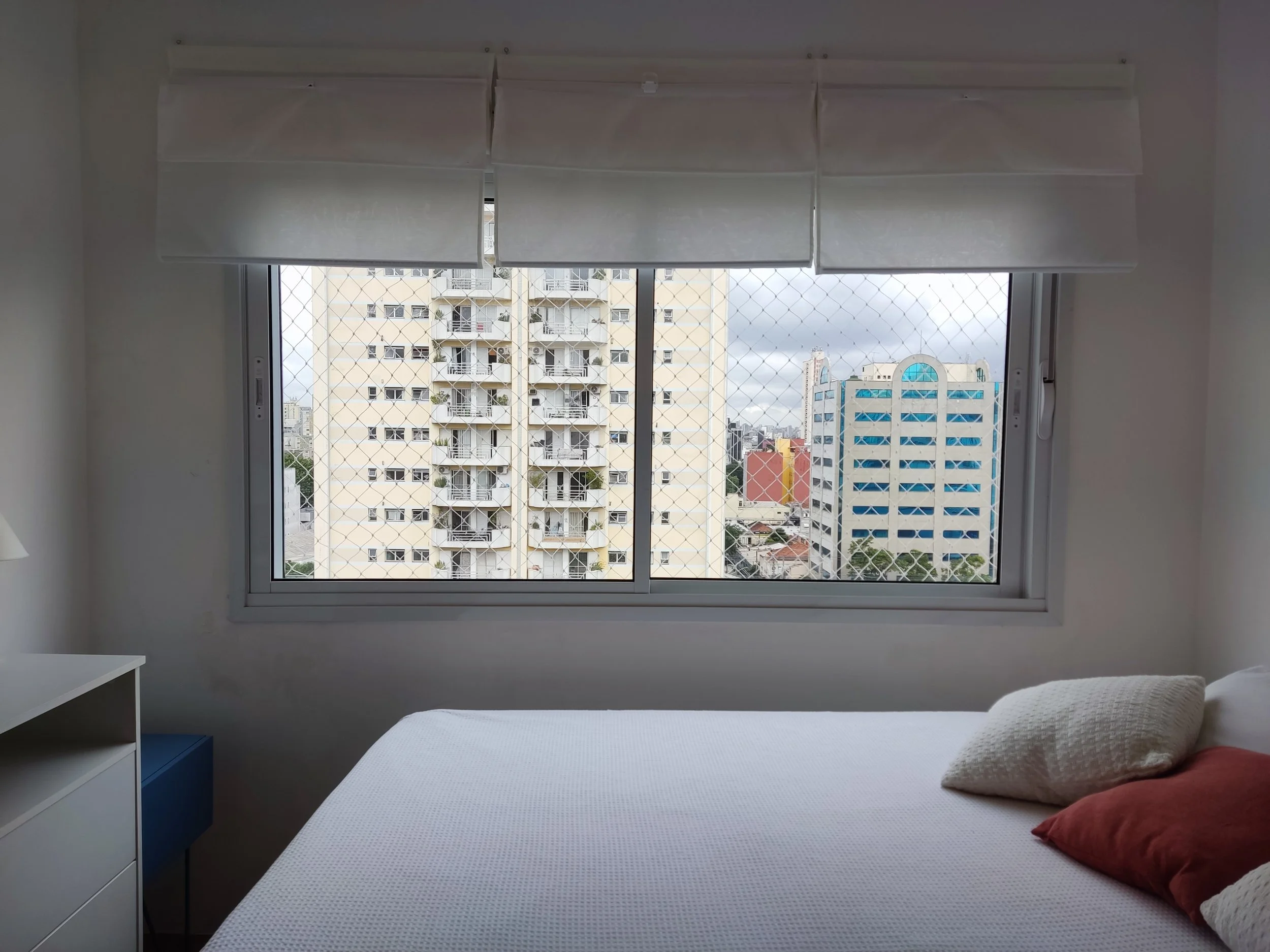 Quarto com uma janela grande com proteção de tela, vista de edifícios residenciais e comerciais, cama com travesseiros brancos e vermelhos, cômoda branca ao lado.