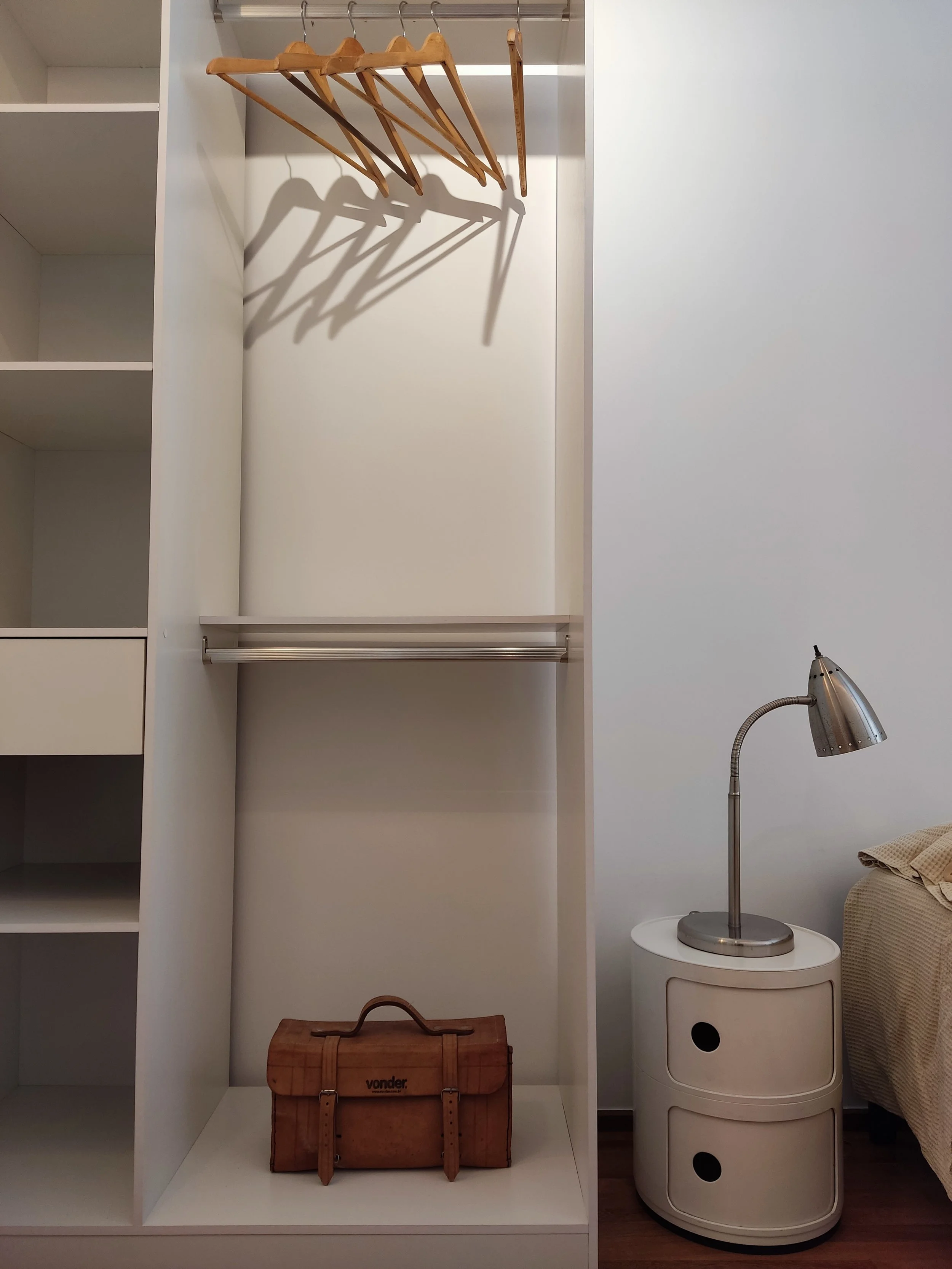 Armário de closet com cabides de madeira na parte superior, uma caixa de ferramentas de madeira na parte inferior, ao lado uma mesa de cabeceira branca com gavetas e luminária de aço