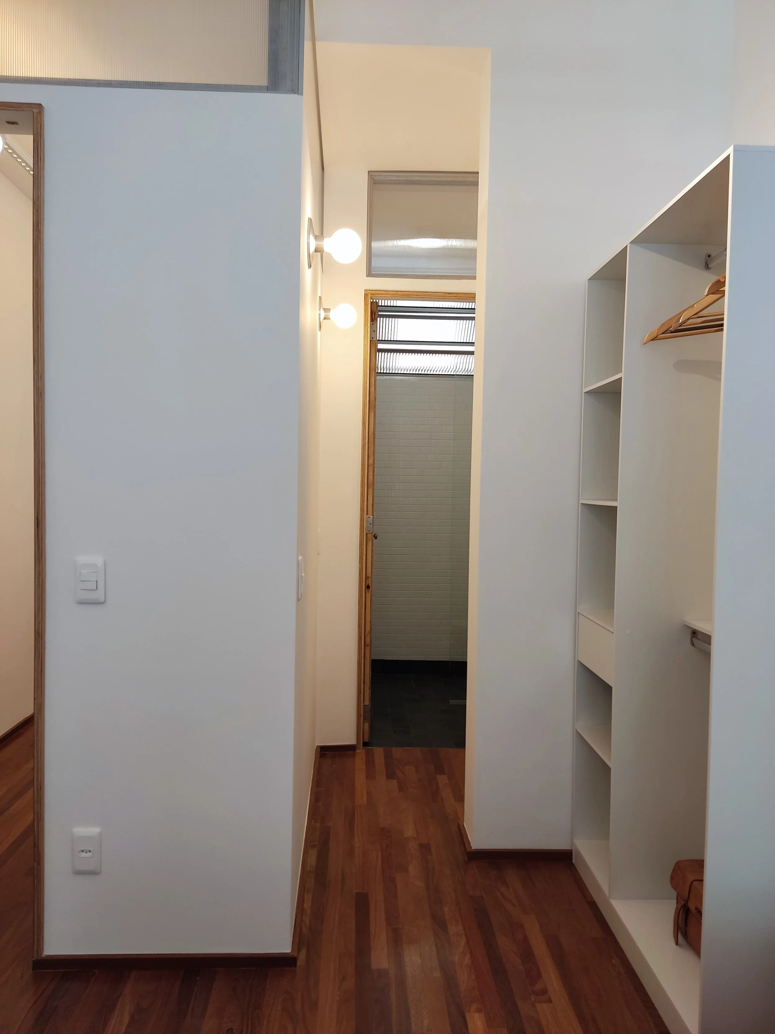 Corredor de uma casa com piso de madeira escura, parede branca, iluminação de duas luminárias na parede, porta de vidro no fundo levando a um espaço com azulejos brancos, e uma roupa de cama de madeira na parede direita.