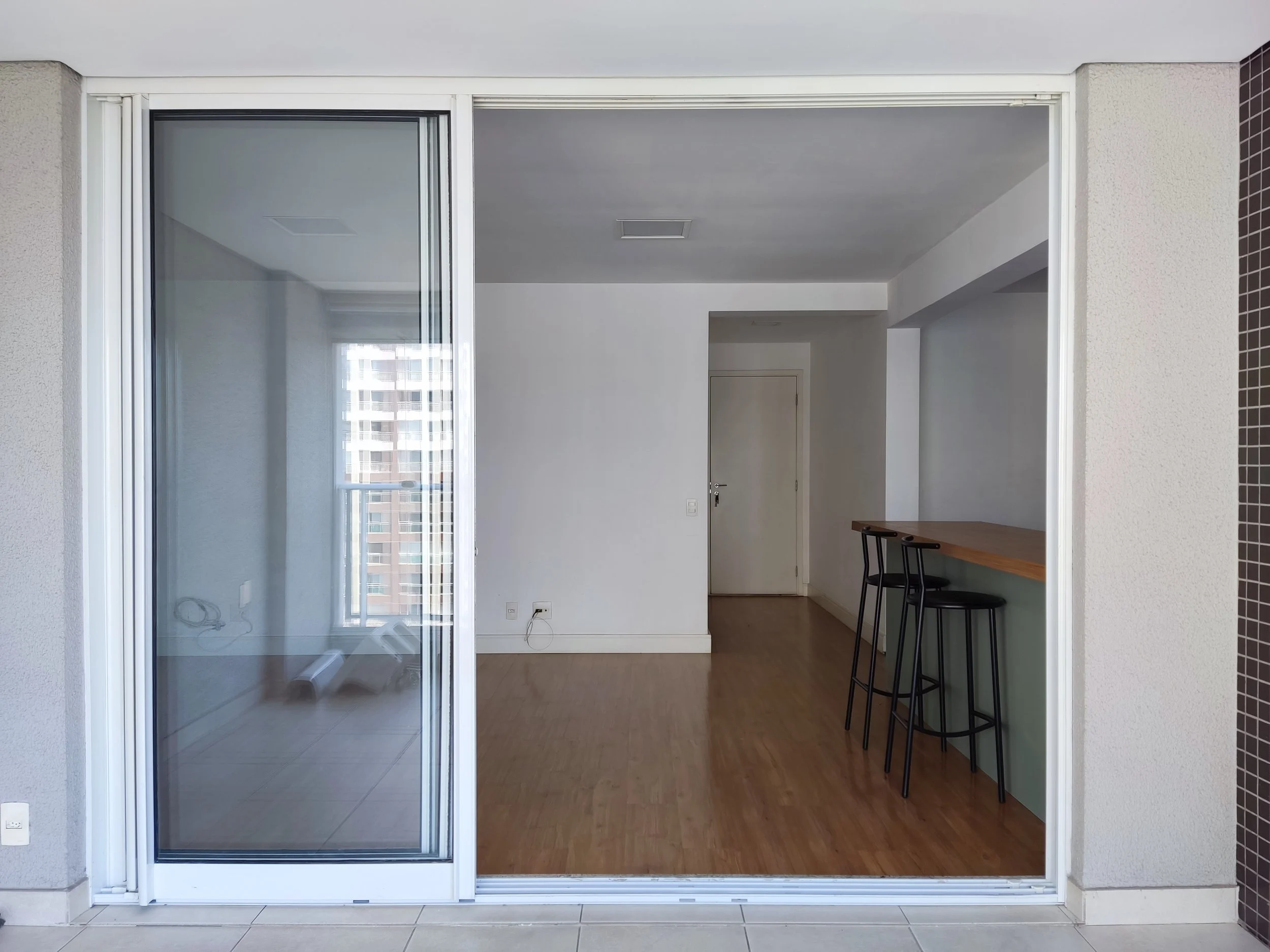 Interior de um apartamento, vista de uma sala com piso de madeira, parede branca, com porta de vidro deslizante que leva a um espaço com varanda, uma porta branca ao fundo, e uma bancada com três bancos altos ao lado direito.