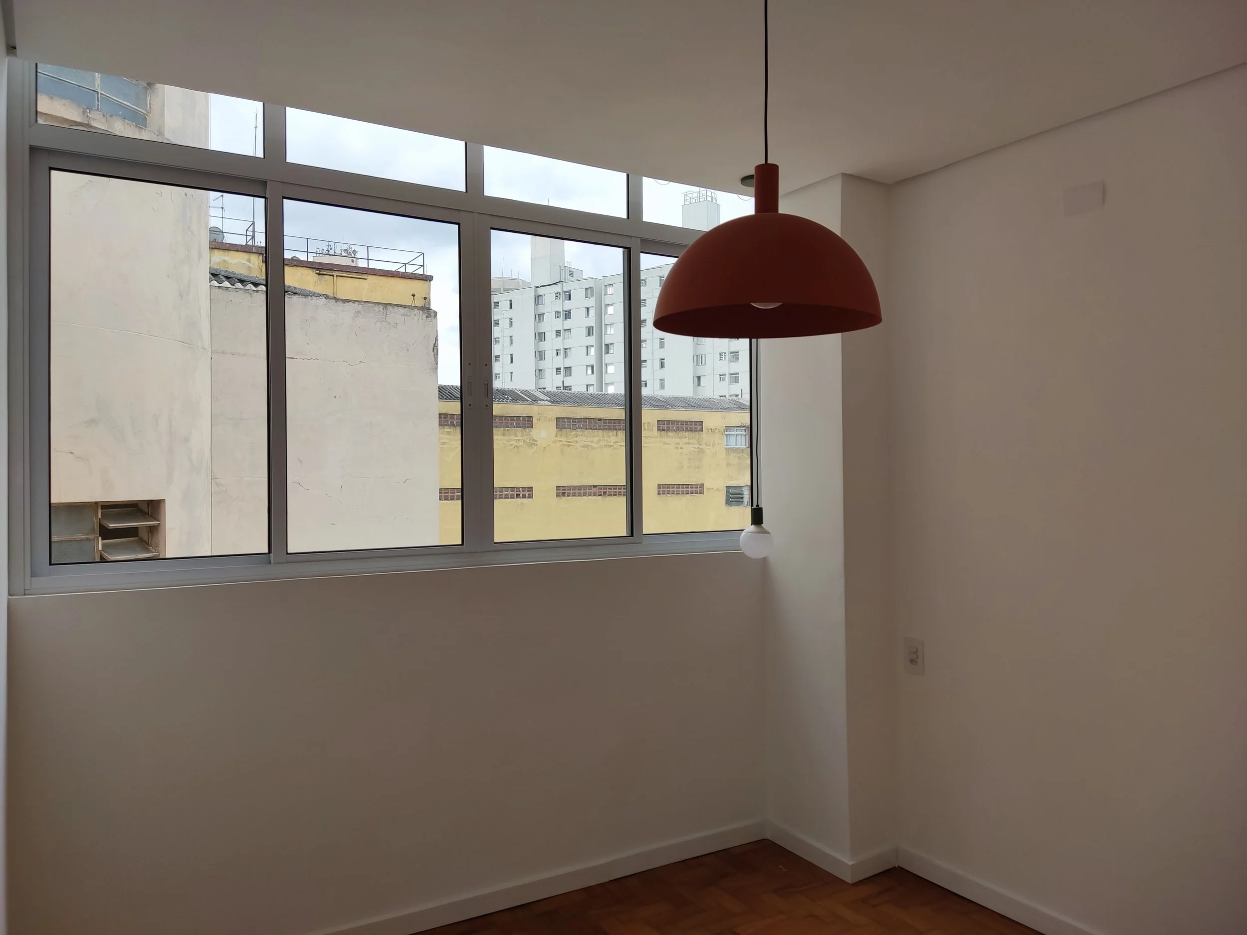 Sala com janela grande, parede branca, luminária pendente vermelha e lâmpada acesa, piso de madeira.