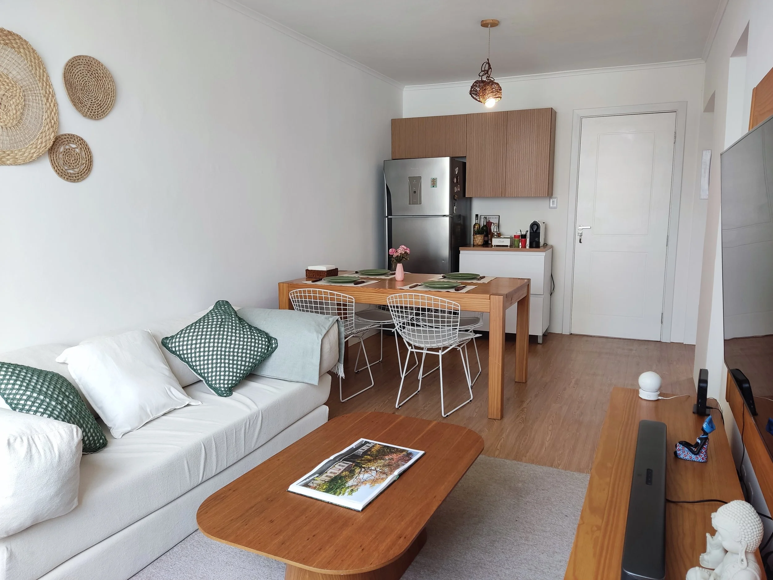 Sala de estar e cozinha em estilo clean com móveis de madeira, sofá branco com almofadas verdes, mesa de centro de madeira, mesa de jantar, cadeiras brancas de metal, geladeira de inox, prateleiras de madeira, televisão, objetos decorativos e piso de