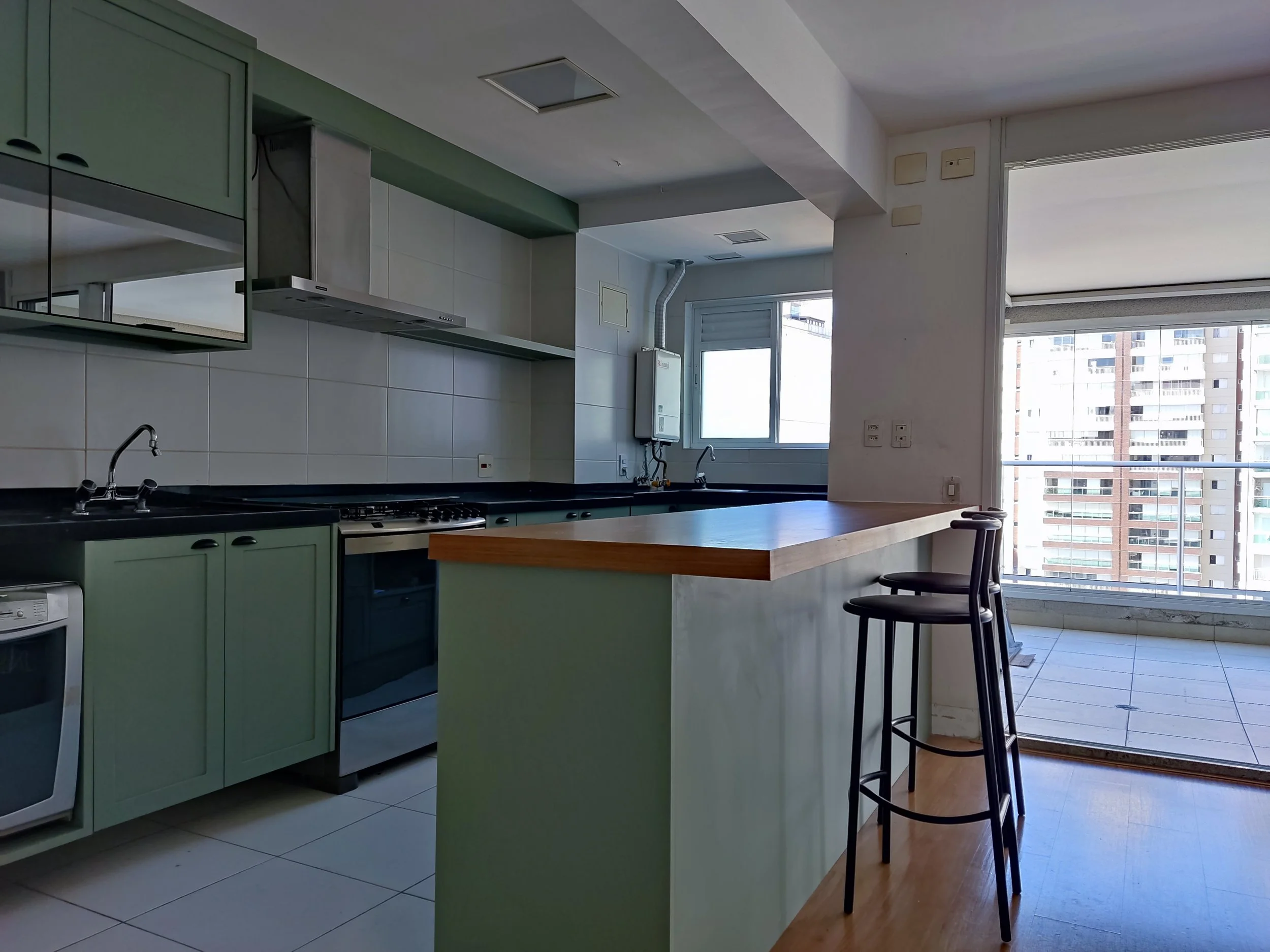 Cozinha moderna com armários verdes, pia dupla, fogão, forno, balcão de madeira, duas banquetas pretas, janela oferecendo vista de prédios altos