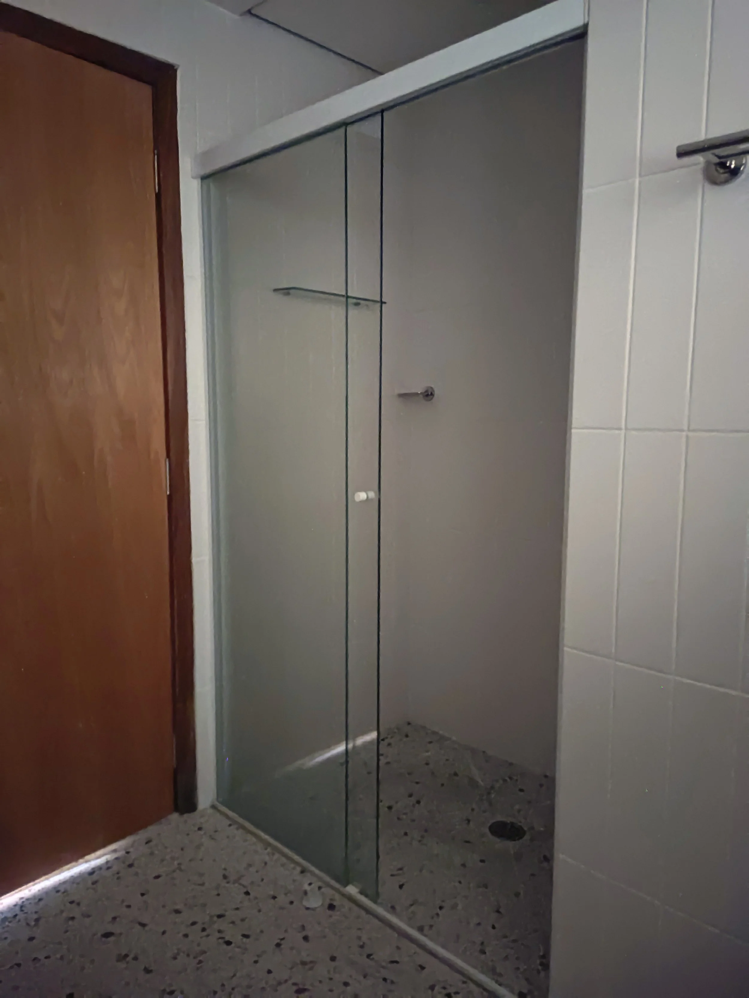 Banheiro com porta de vidro separando o chuveiro, paredes brancas e chão de mármore com detalhes escuros.