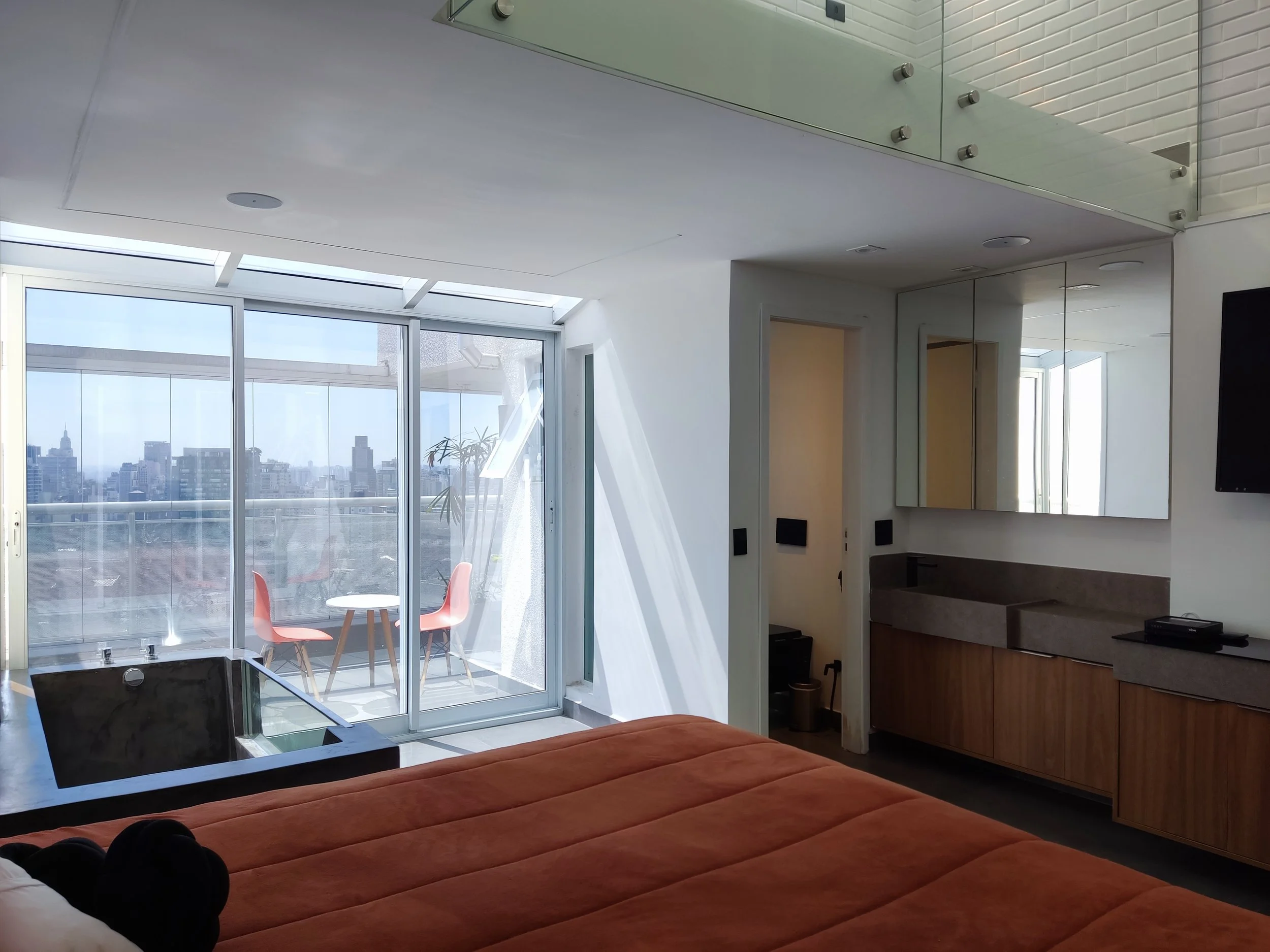 Quarto com cama de tecido laranja, varanda com mesa e cadeiras rosas, janelas amplas com vista para a cidade, espelho grande na parede, televisão