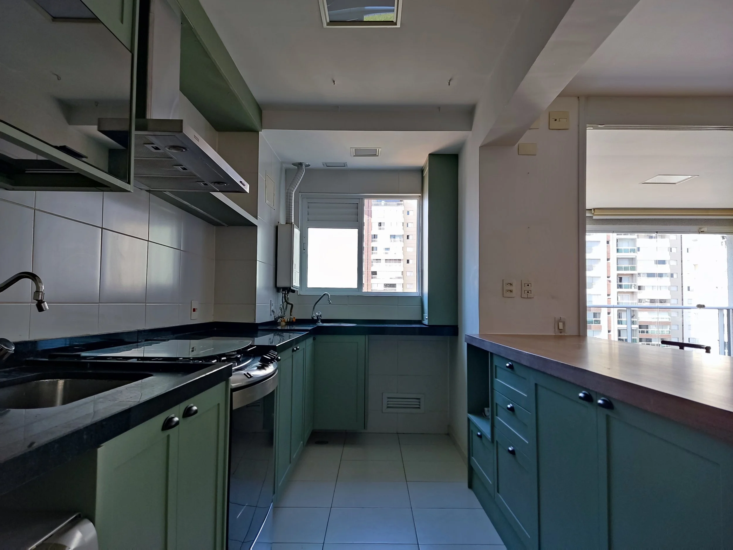 Cozinha de apartamento com armários verdes, pia de aço, cooktop e forno embutido, janela ao fundo com vista de prédios altos, e área de serviço ao lado.