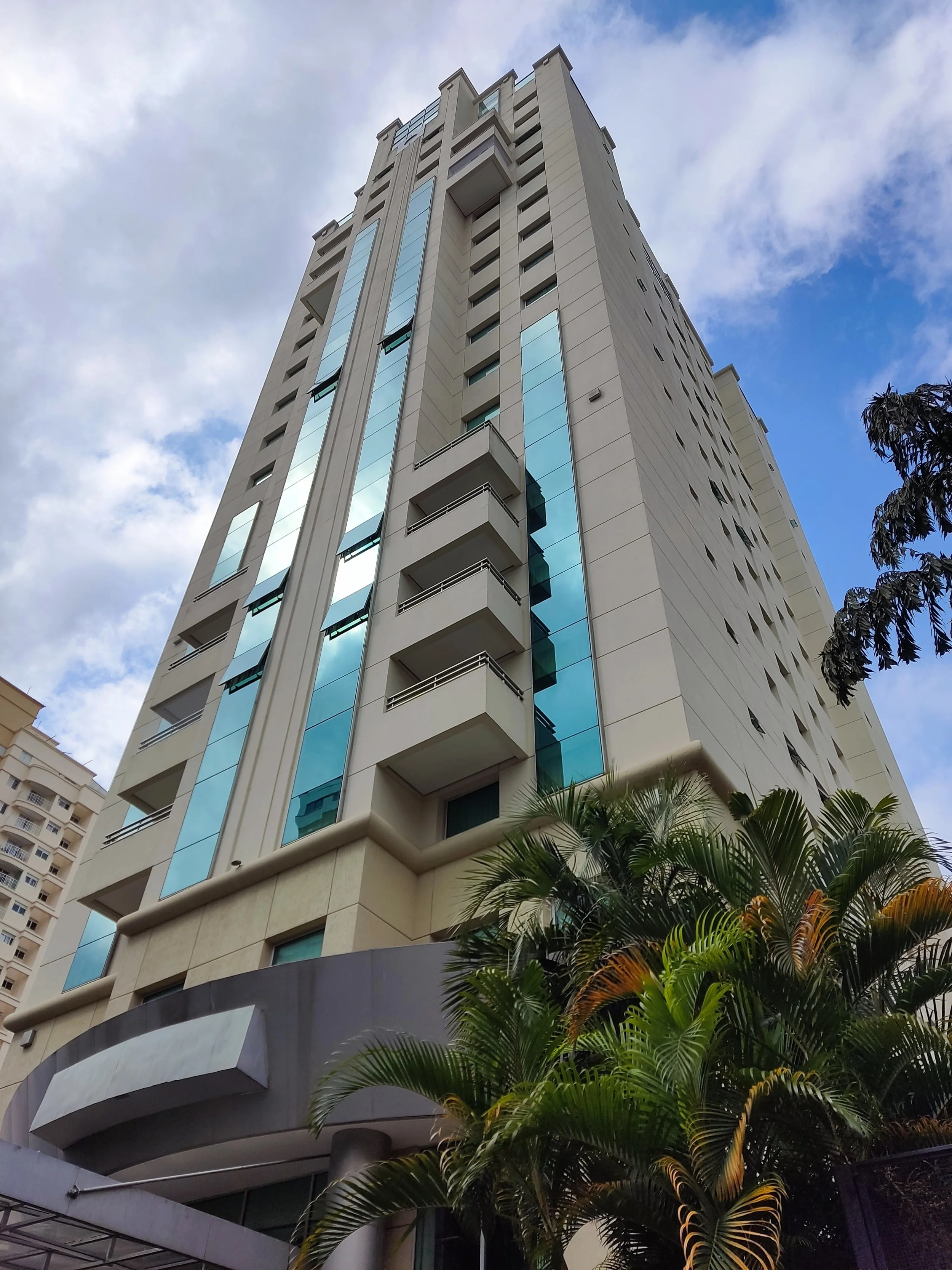 Prédio moderno de altura elevada com janelas de vidro azuis refletoras, cercado por árvores de folhagem verde na frente, sob céu parcialmente nublado.