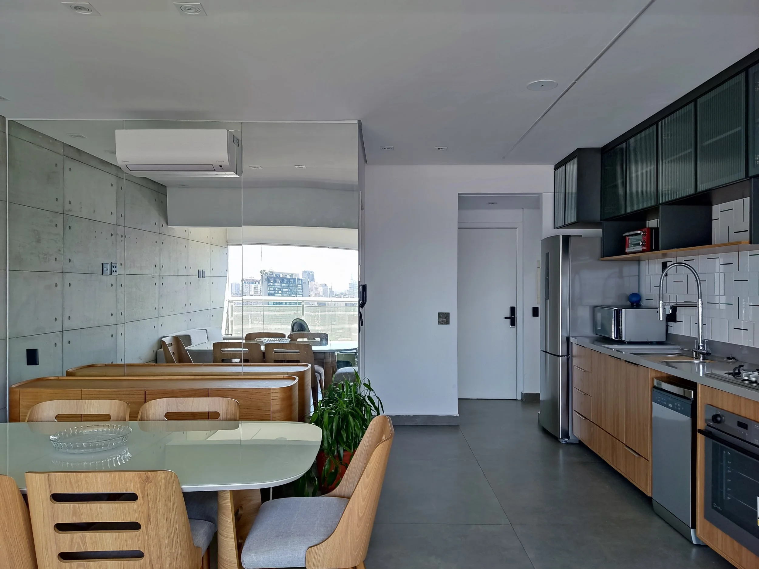 Cozinha moderna com balcão de madeira, eletrodomésticos de aço inox, paredes de azulejos brancos, armários cinza escuro, mesa de jantar de vidro com cadeiras de madeira, vista para uma cidade via janela grande, ar-condicionado no teto.