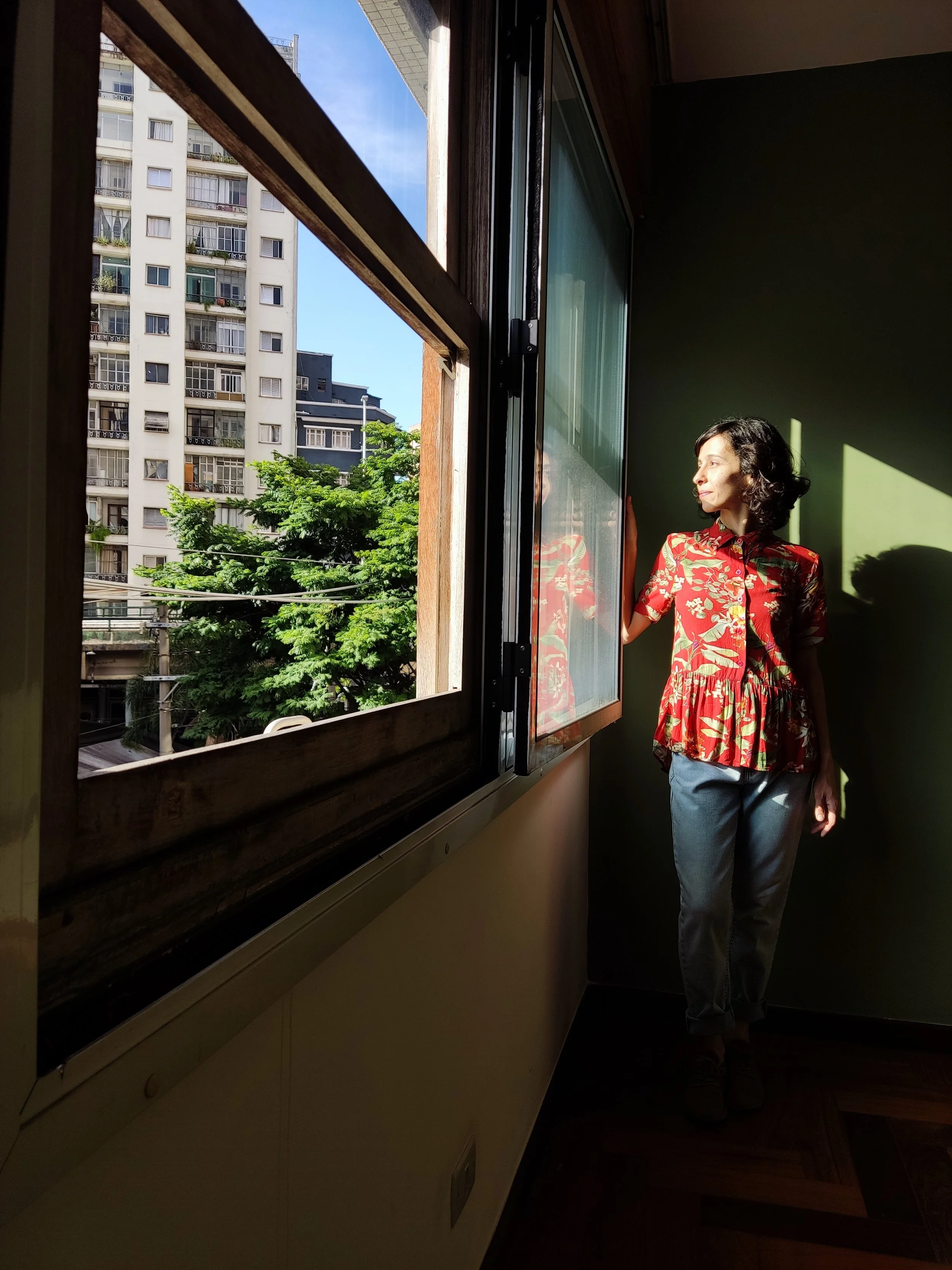 Mulher de cabelos cacheados, usando blusa vermelha com estampa de flores, de pé ao lado de uma janela, olhando para fora, com luz do sol iluminando seu rosto.