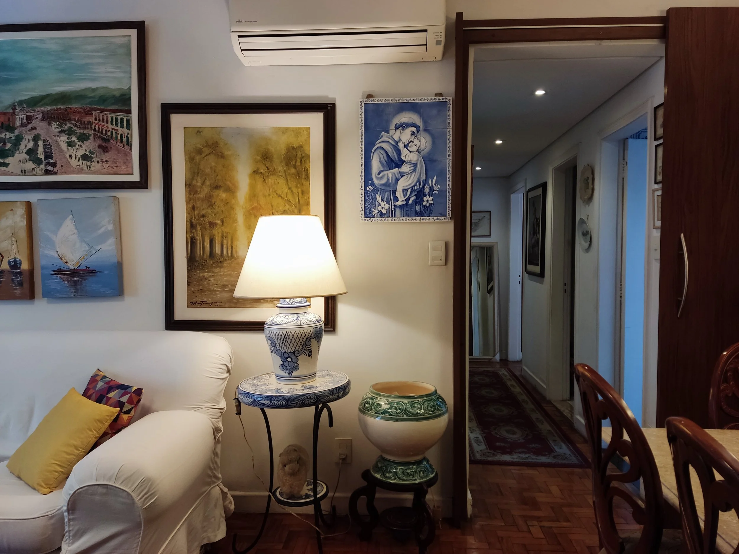 Sala de estar decorada com quadros na parede, uma luminária azul e branca em uma mesinha, uma estante com um vaso decorativo, sofá branco com almofadas coloridas, e uma porta que leva a um corredor.