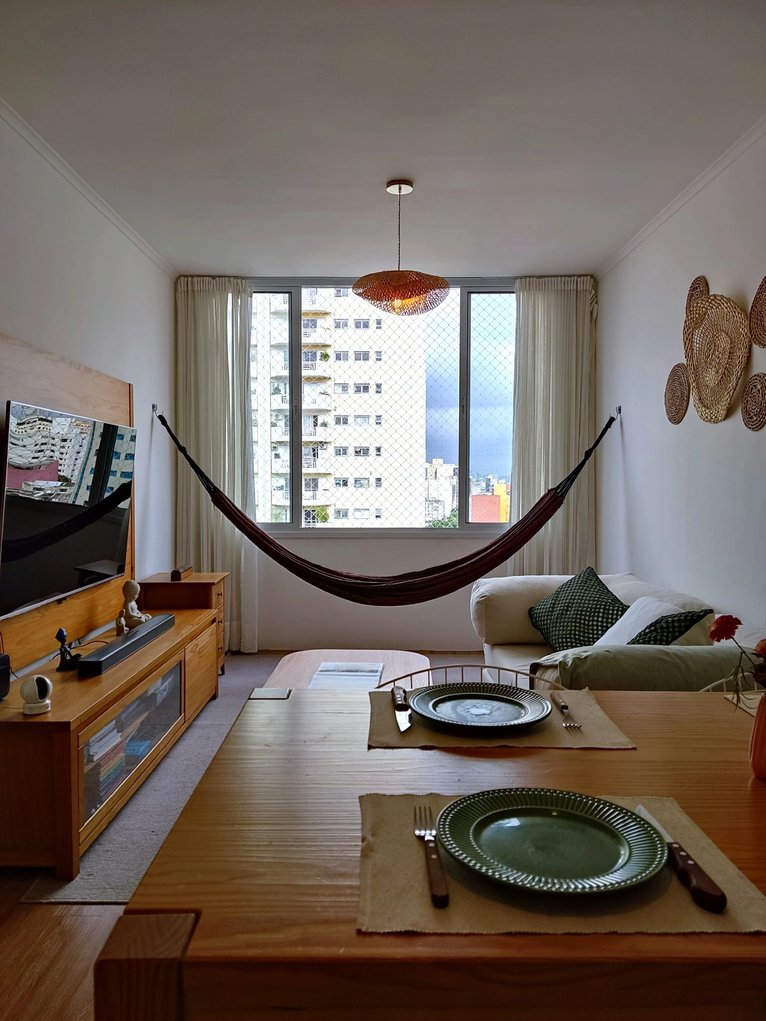 Sala de estar com mesa de jantar, sofá branco com almofadas verdes, rede de balanço na janela, televiso, decoração de palha na parede, janelas com cortinas brancas, iluminação natural.