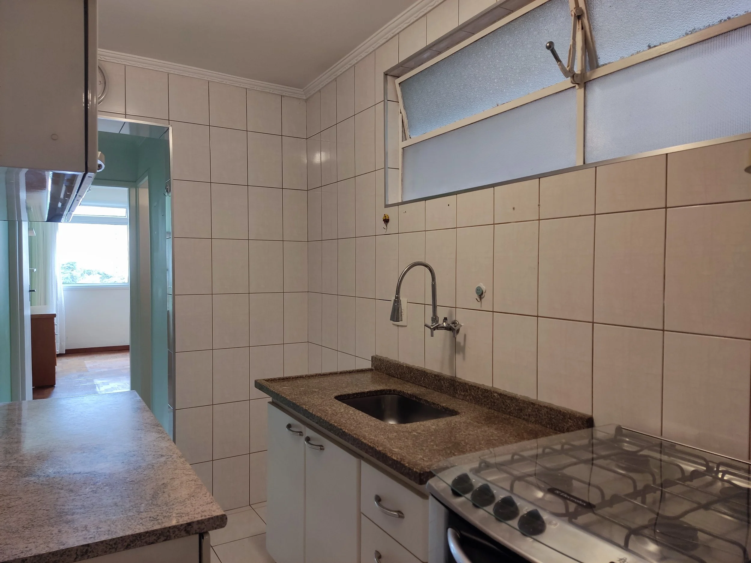 Cozinha com pia de granito, fogão e parede de azulejos, janela grande acima da pia, porta levando à sala.