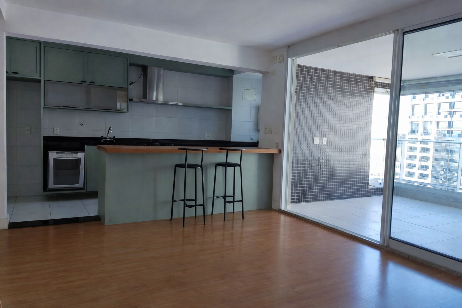 Sala de estar com cozinha americana, piso de madeira, bancada com duas cadeiras, área de sacada com vista para prédios