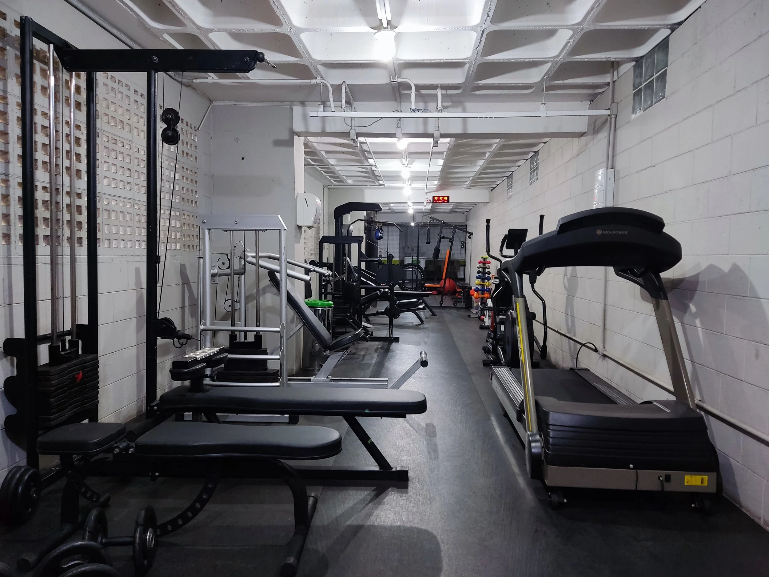 Sala de musculação com equipamentos de ginástica, incluindo esteira, peso livre, banco de supino, e máquinas de treinamento, em ambiente com paredes de blocos de vidro e piso de borracha