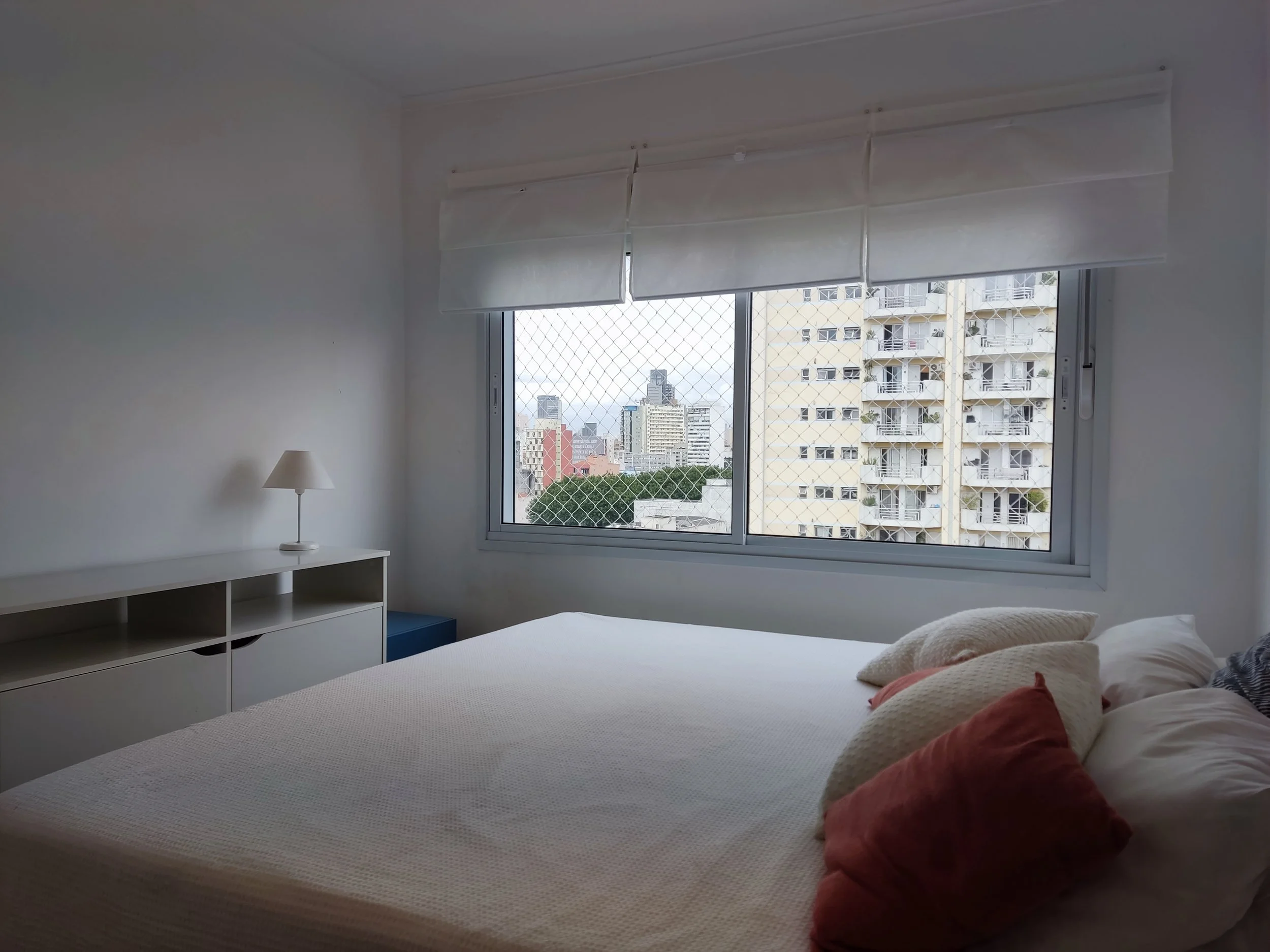 Quarto branco com cama com travesseiros e janelas grandes com vista para edifícios urbanos.
