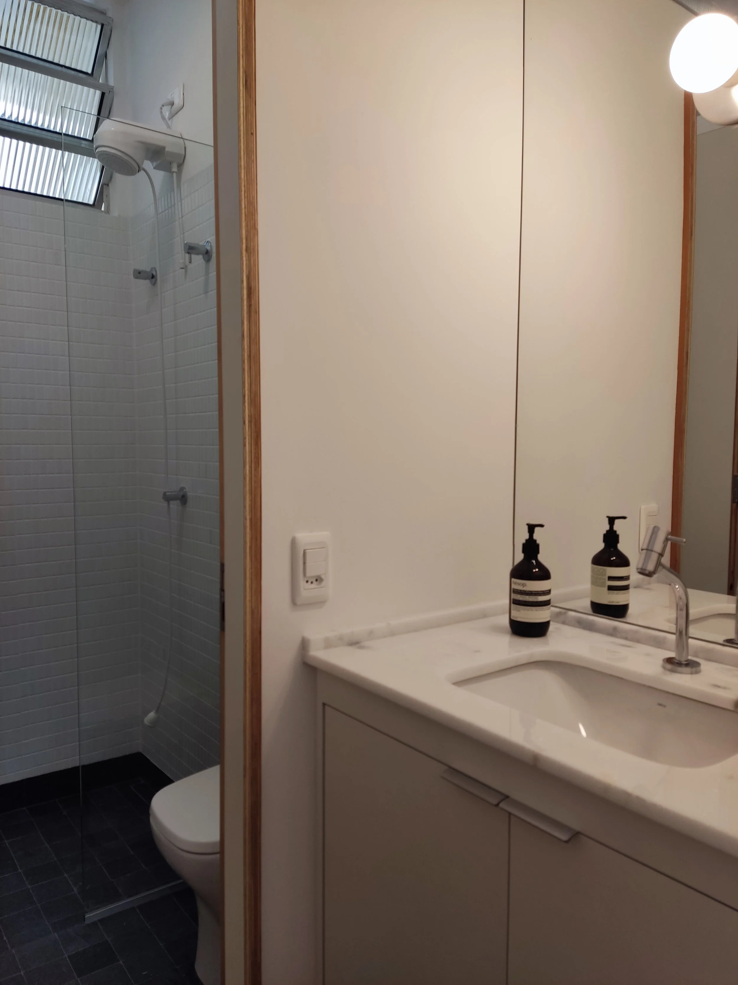 Banheiro com pia de mármore, dois dispensers de sabonete, espelho grande, armário inferior, parede branca e ducha com janela de vidro na área do chuveiro.