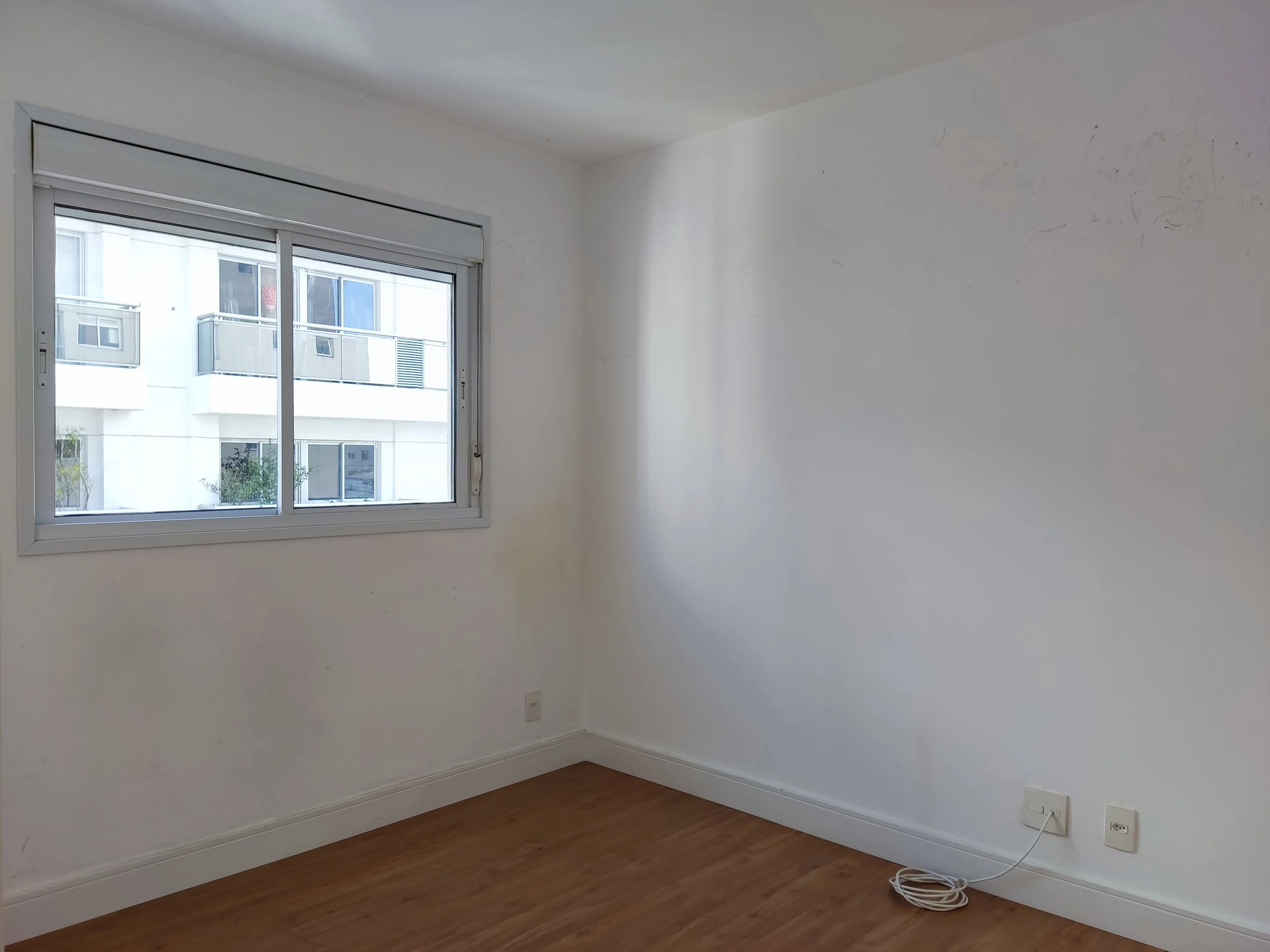 Quarto vazio com janela, piso de madeira, parede branca e tomadas elétricas.