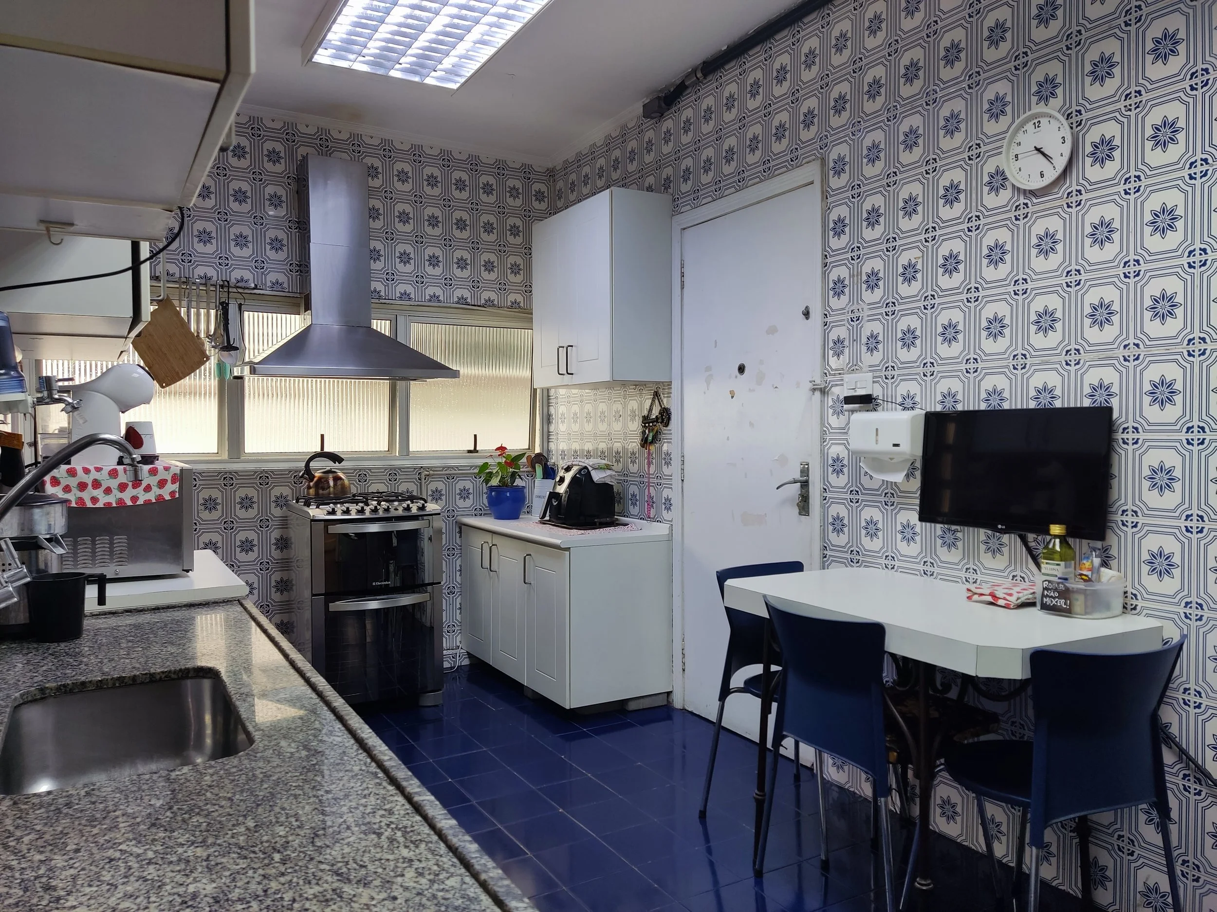 Cozinha decorada com paredes revestidas em azulejos azuis e brancos, com área de pia, fogão a gás, mesa com quatro cadeiras azuis, televisão na parede e janela com cortina de vidro.