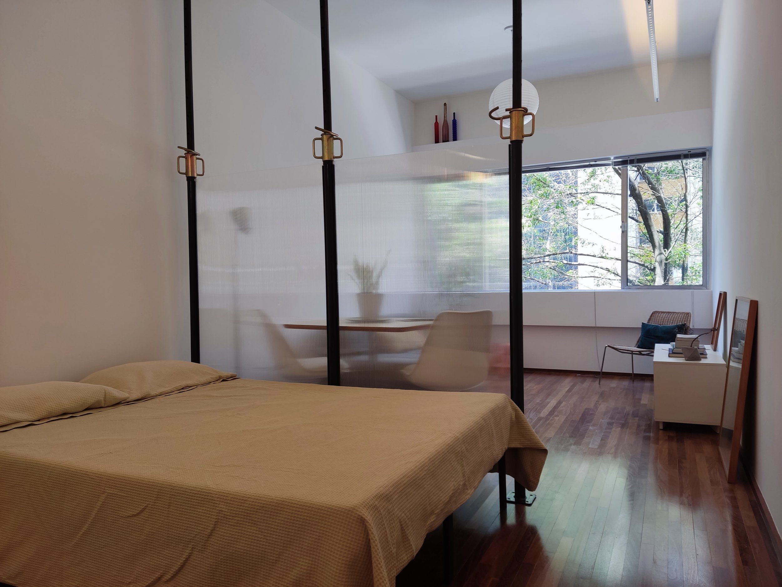 Quarto com cama king size, divisória de ambiente com cortina de malha transparente, mesa de jantar com cadeiras na área de estar, grande janela com vista para árvores, piso de madeira, decoração minimalista com algumas obras de arte e objetos de deco