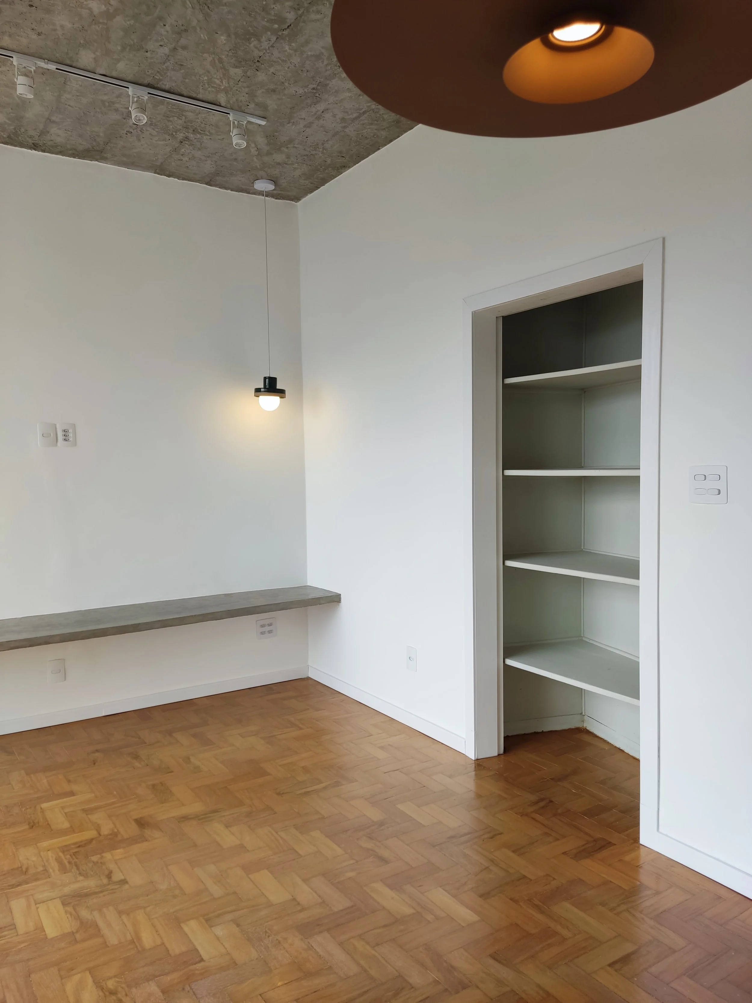 Quarto com piso de parquet, parede branca, prateleiras embutidas brancas, iluminação com lâmpada pendurada e teto de concreto aparente.