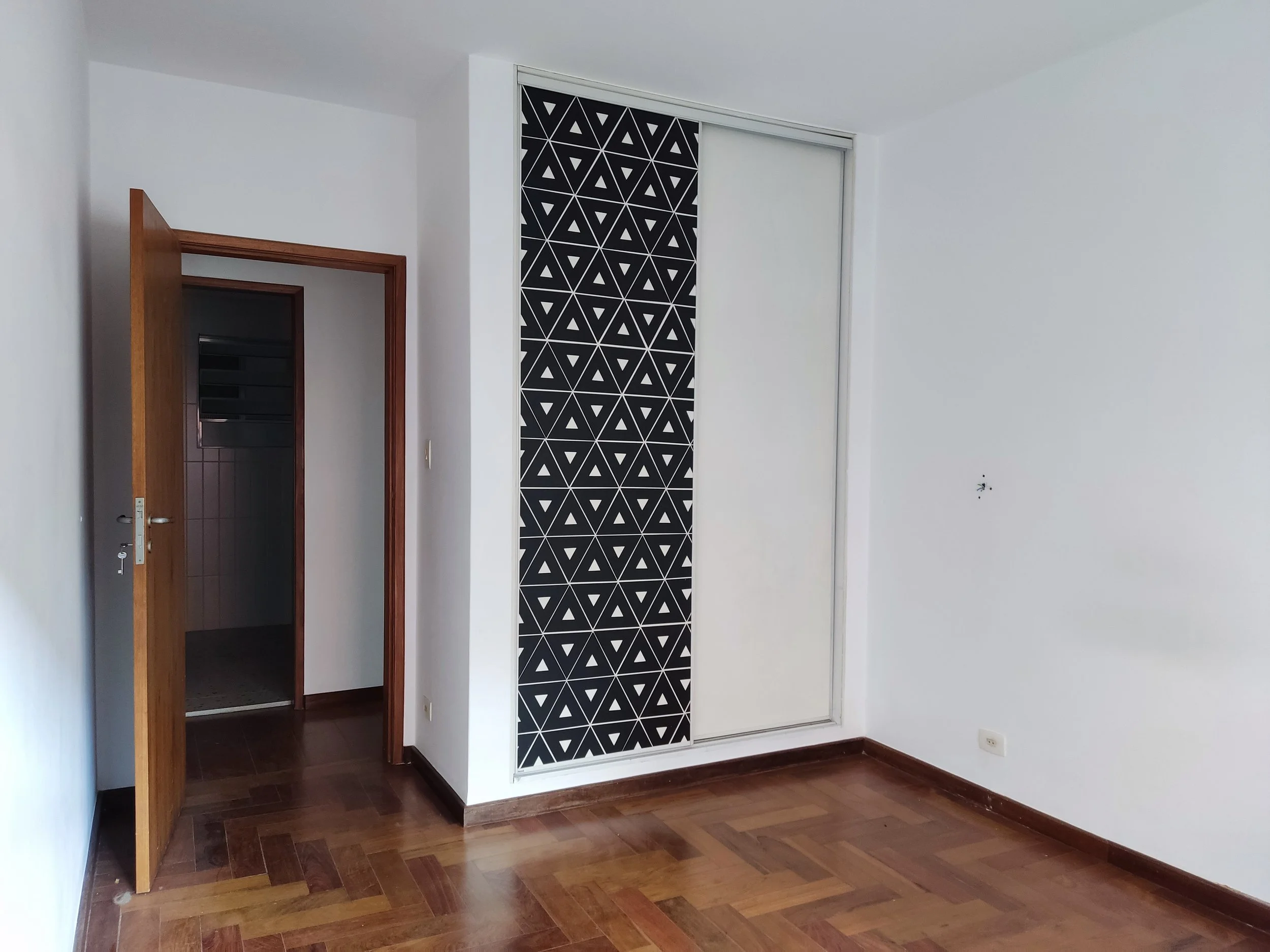 Quarto vazio com piso de madeira, parede branca, porta de madeira aberta e armário de correr com portas brancas e uma de painel preto com padrão geométrico. 