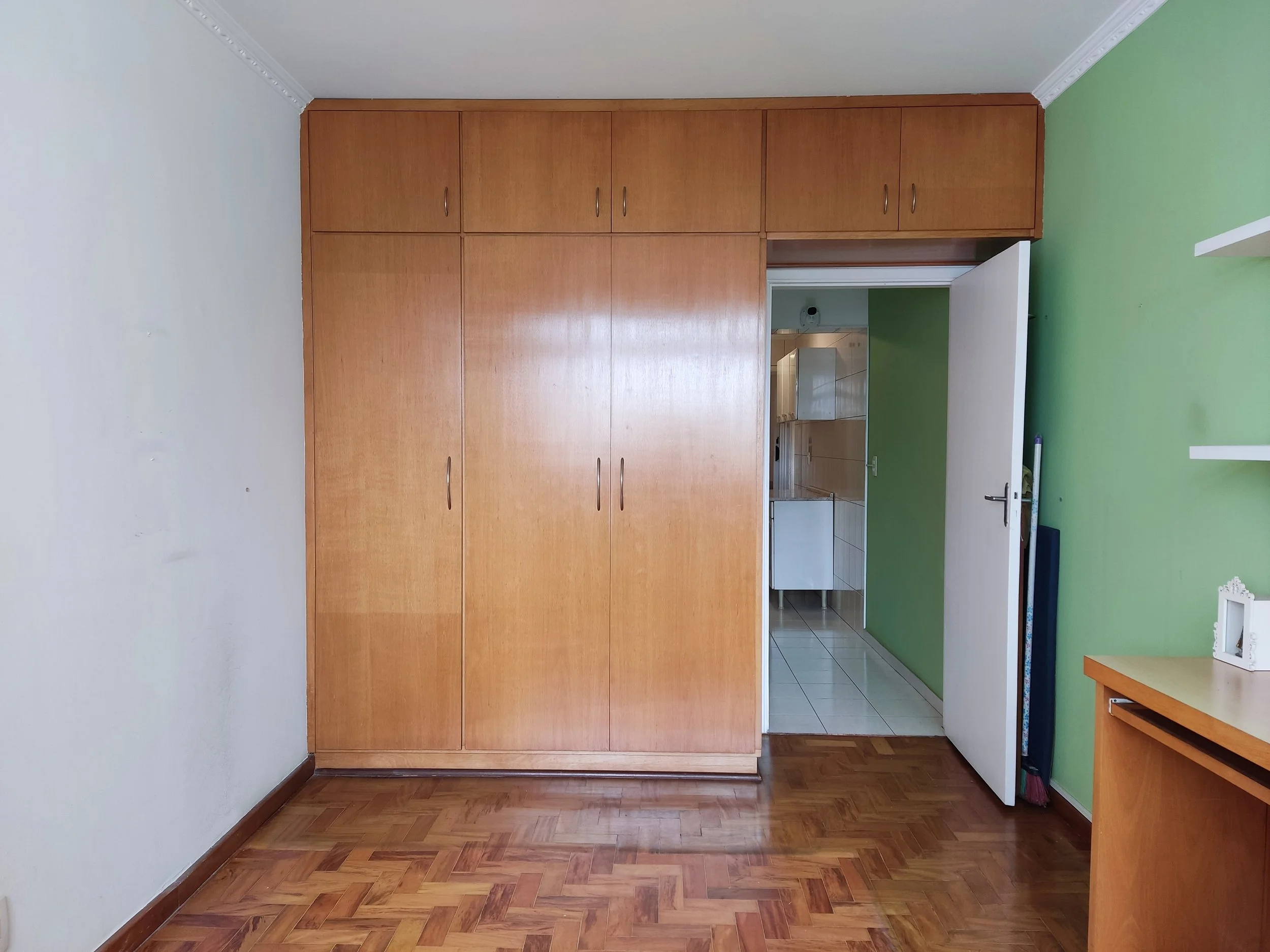 Quarto com armário de madeira e parede verde, porta aberta para a cozinha com piso branco e paredes brancas.