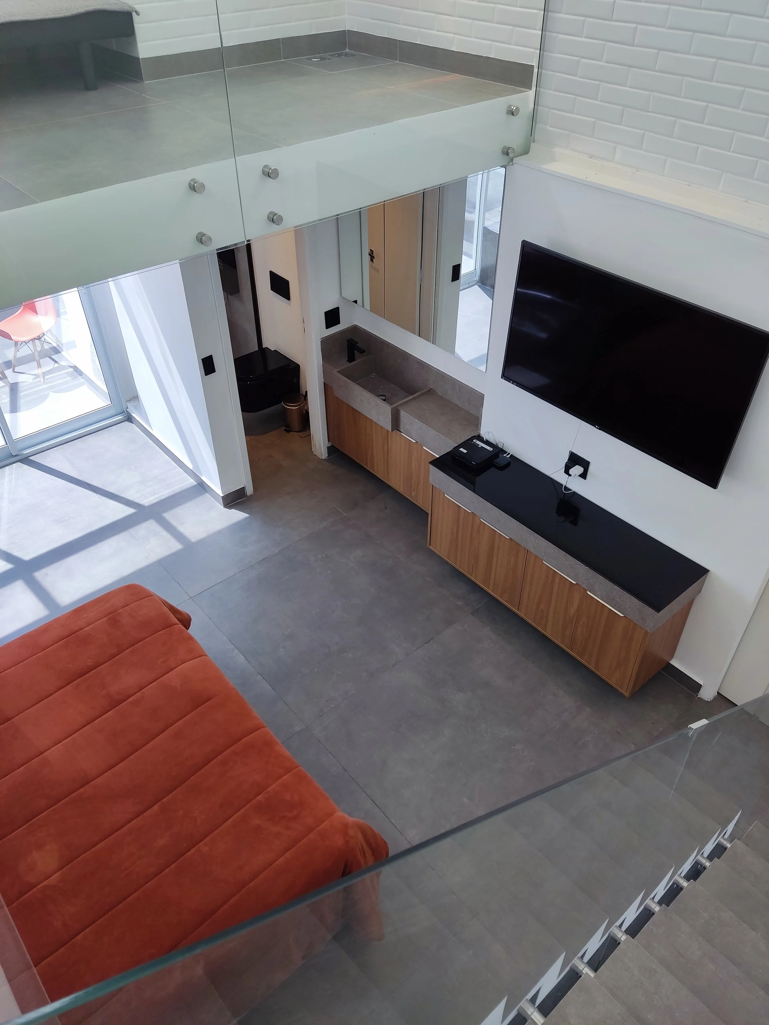 Visão do interior de um apartamento que inclui um sofá laranja, uma parede com televisão de tela grande, uma bancada moderna com pia, uma parede de tijolos brancos e uma escada de vidro com corrimão de metal, além de uma porta de vidro que dá acesso 