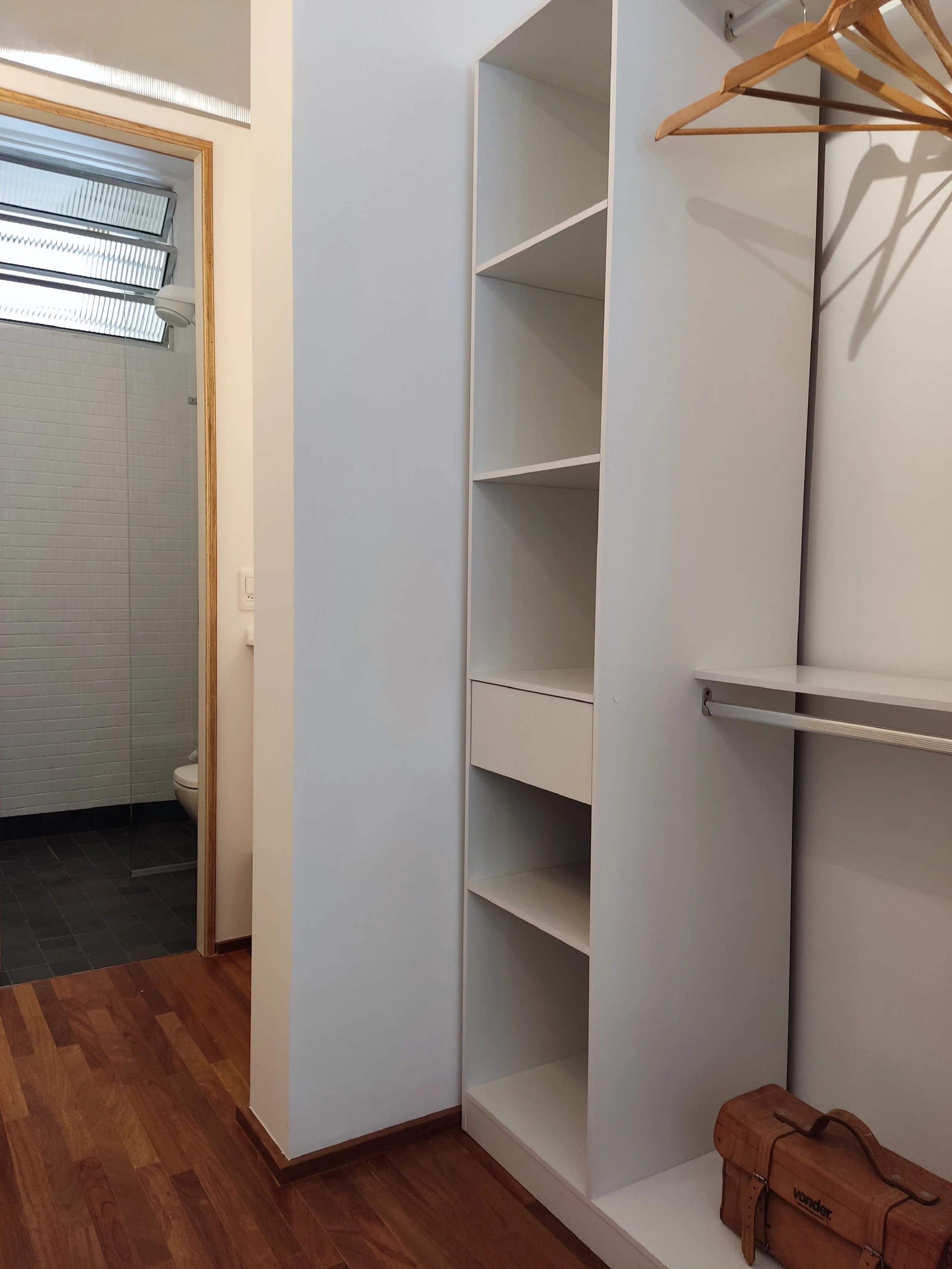 Armário de parede branco com prateleiras e uma gaveta, ao lado de um espaço vazio para cabides, no interior de um quarto. Ao fundo, uma porta leva ao banheiro com piso de azulejos escuros e uma pia.